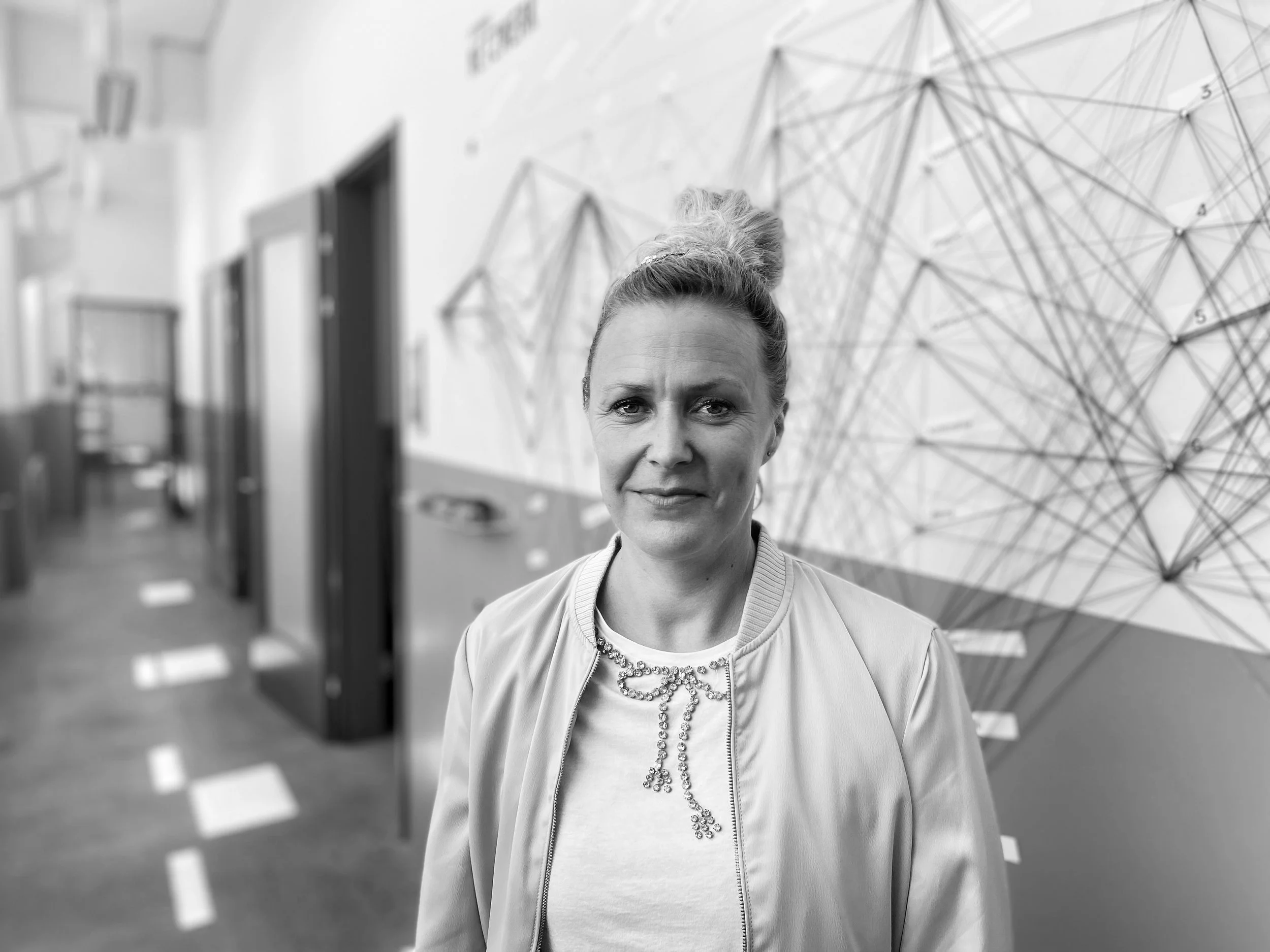 Schwarz-weiß-Fotografie einer Frau mit aufgestyltem Haar, die vor einer Wand mit geometrischem Kunstwerk steht.