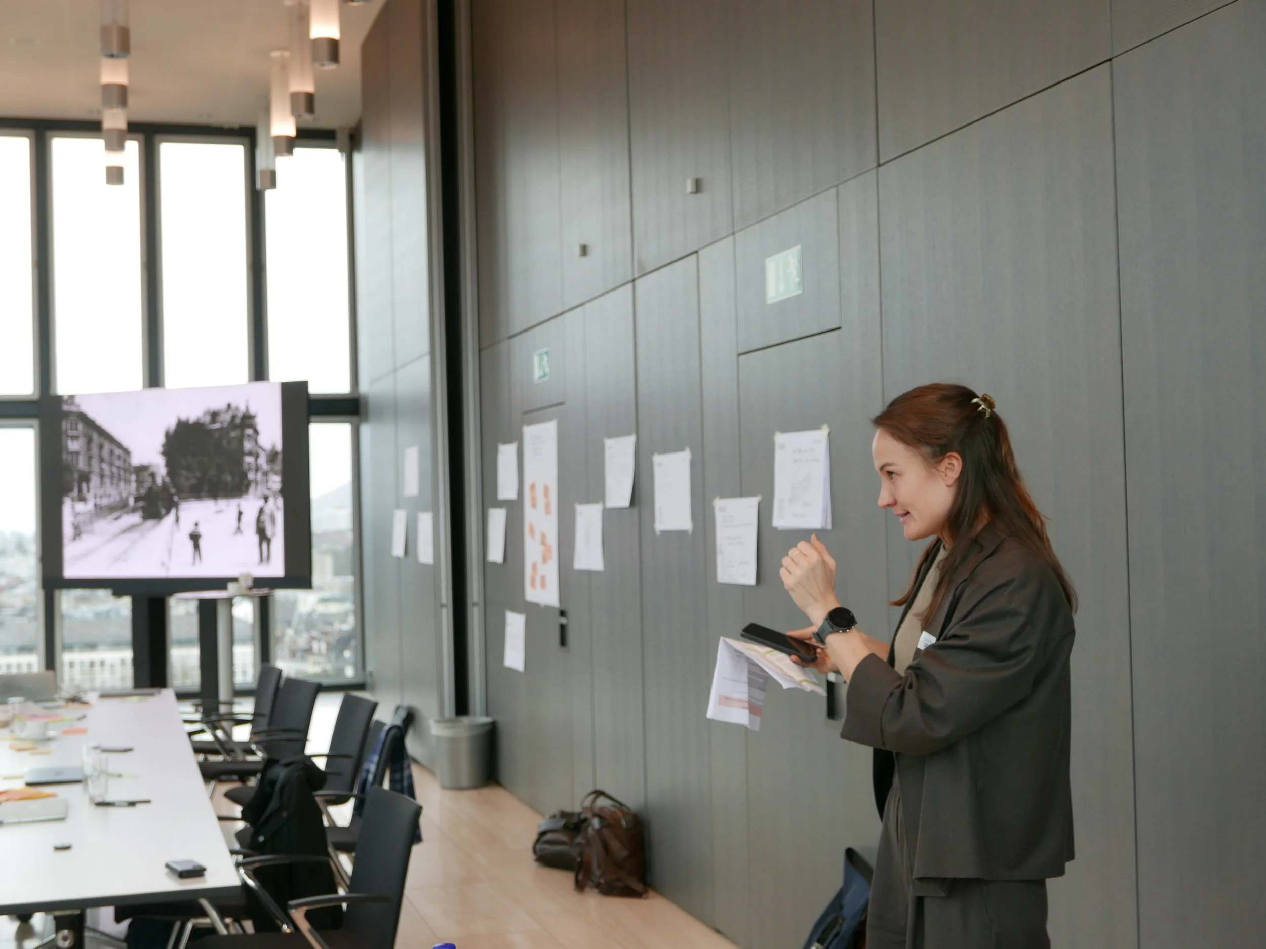 Frau in Businesskleidung, die in einem modernen Konferenzraum steht, während sie mit einem Notizbuch in der Hand spricht. Im Raum gibt es eine lange Tafel, Stühle, große Fenster, eine Präsentationswand mit Fotos und Notizen sowie einige Gegenstände auf dem Tisch.