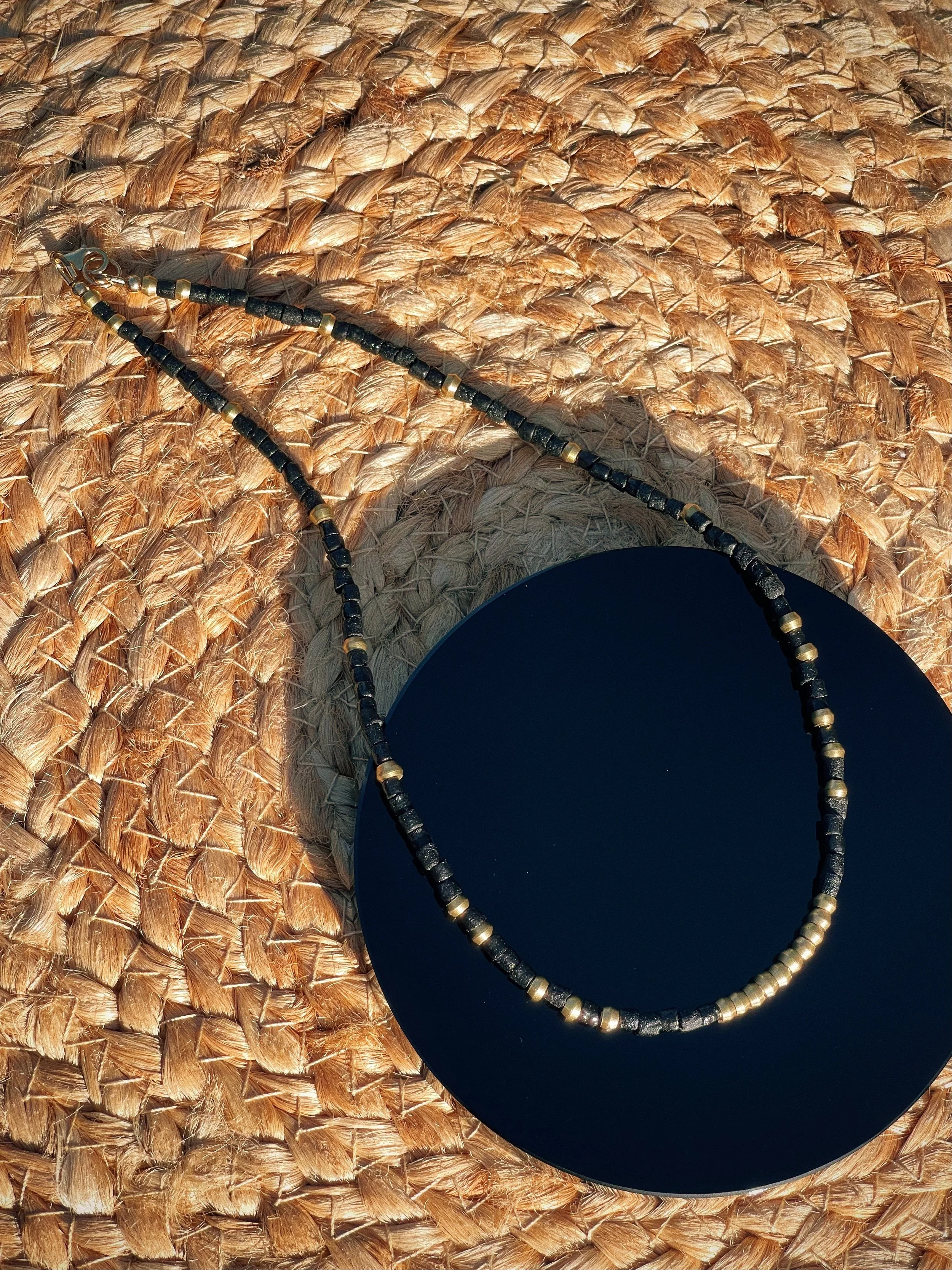Undertow handmade beaded necklace by Tempered Earth, featuring black sandcast beads and brass accents on an onyx slab and woven natural-fiber surface.