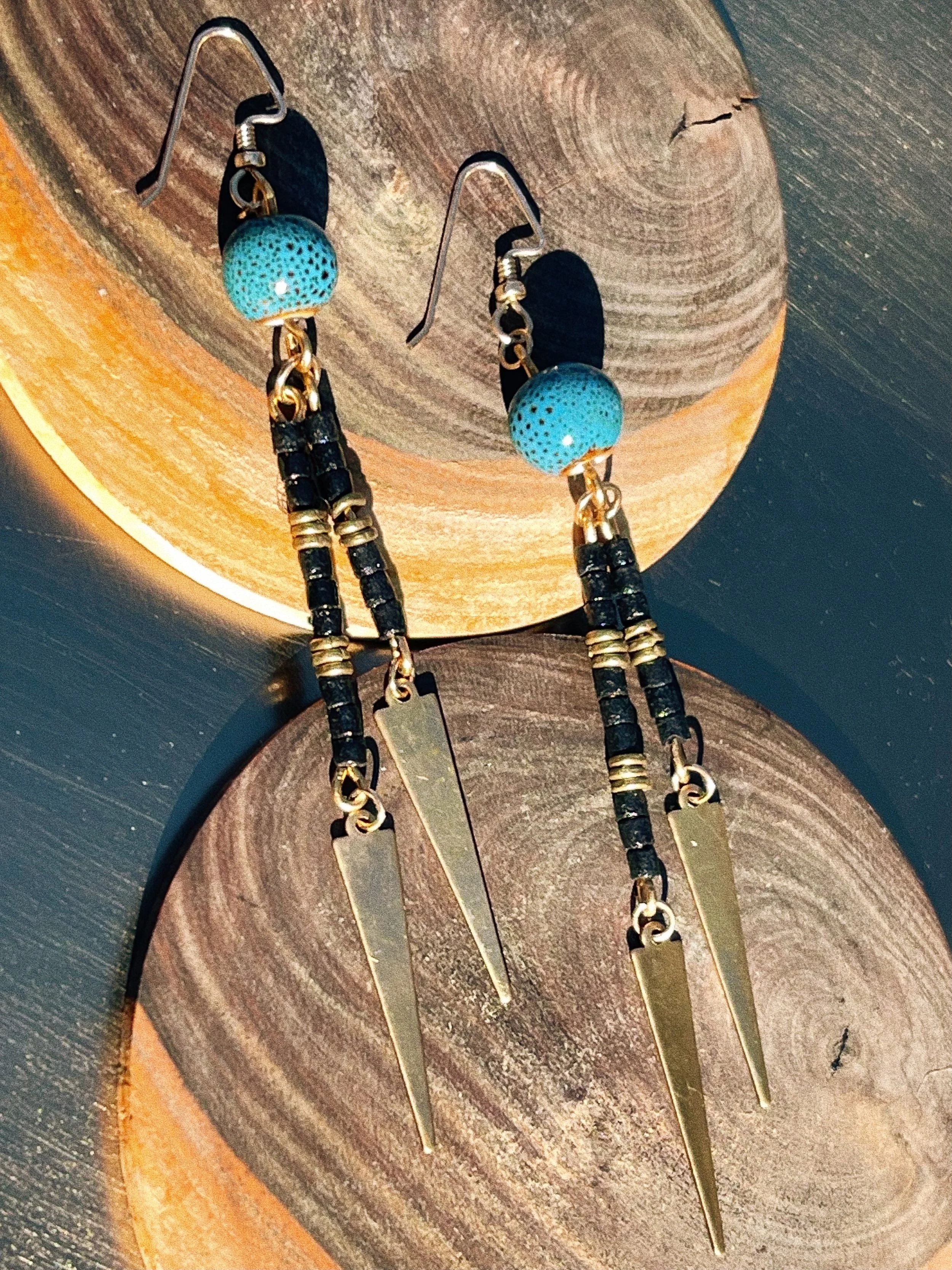 Gaia handmade earrings by Tempered Earth, featuring black sandcast beads, brass accents, and blue ceramic beads, displayed on a gold dish.
