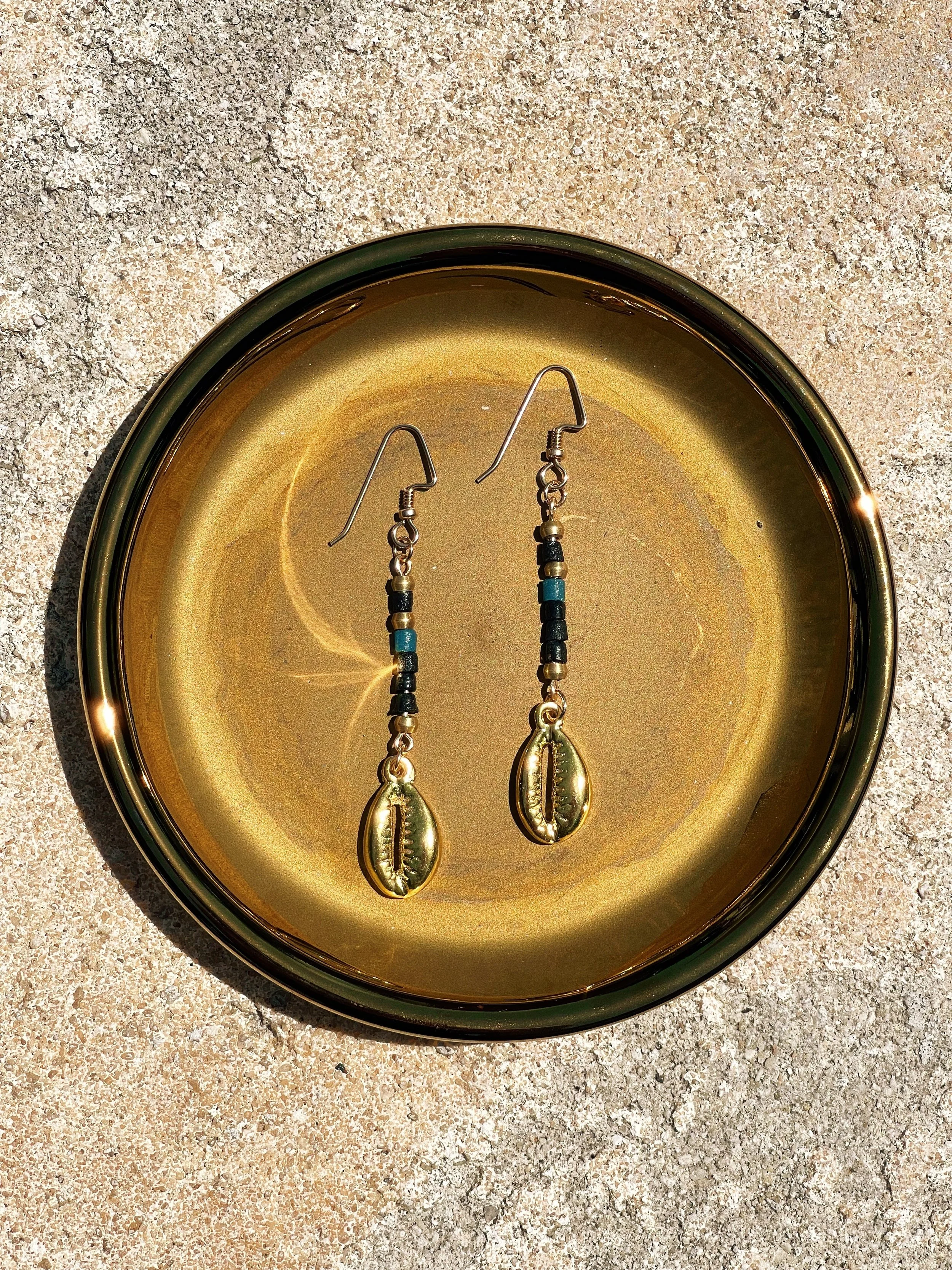 A pair of handmade beaded earrings with cowrie shell pendants resting in a round, shiny, gold-colored dish on a sandy surface.