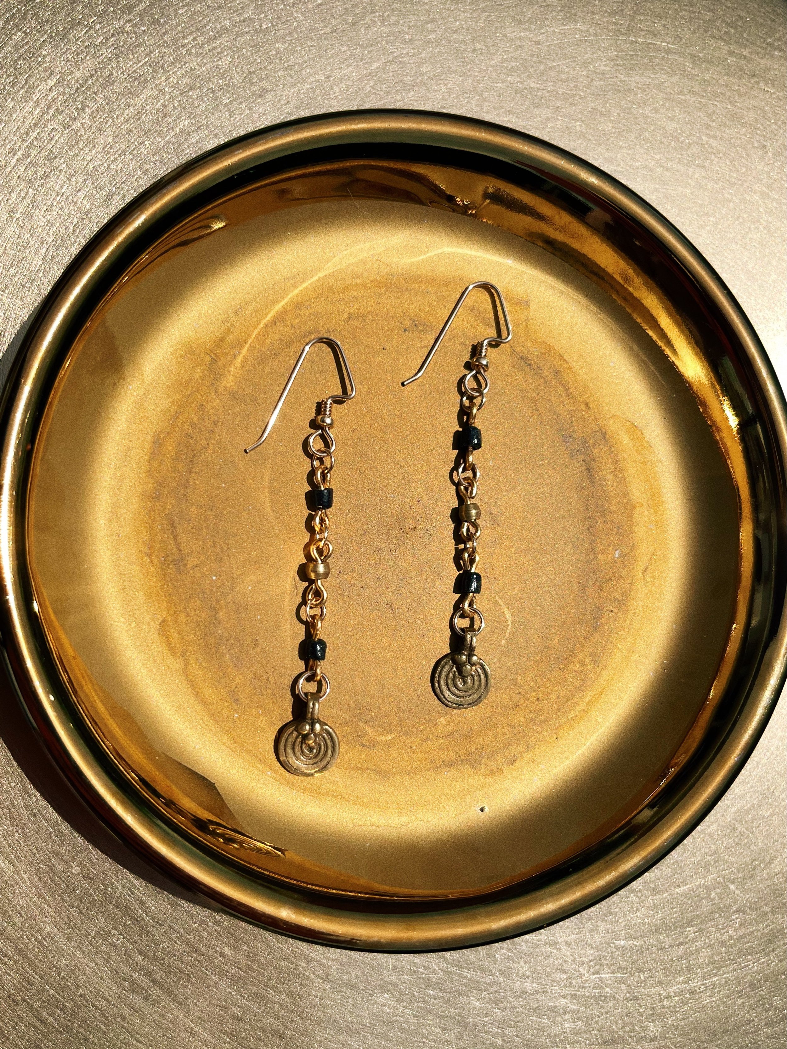 Relic handmade earrings by Tempered Earth, featuring black beads and circular brass pendants, photographed in a gold dish.