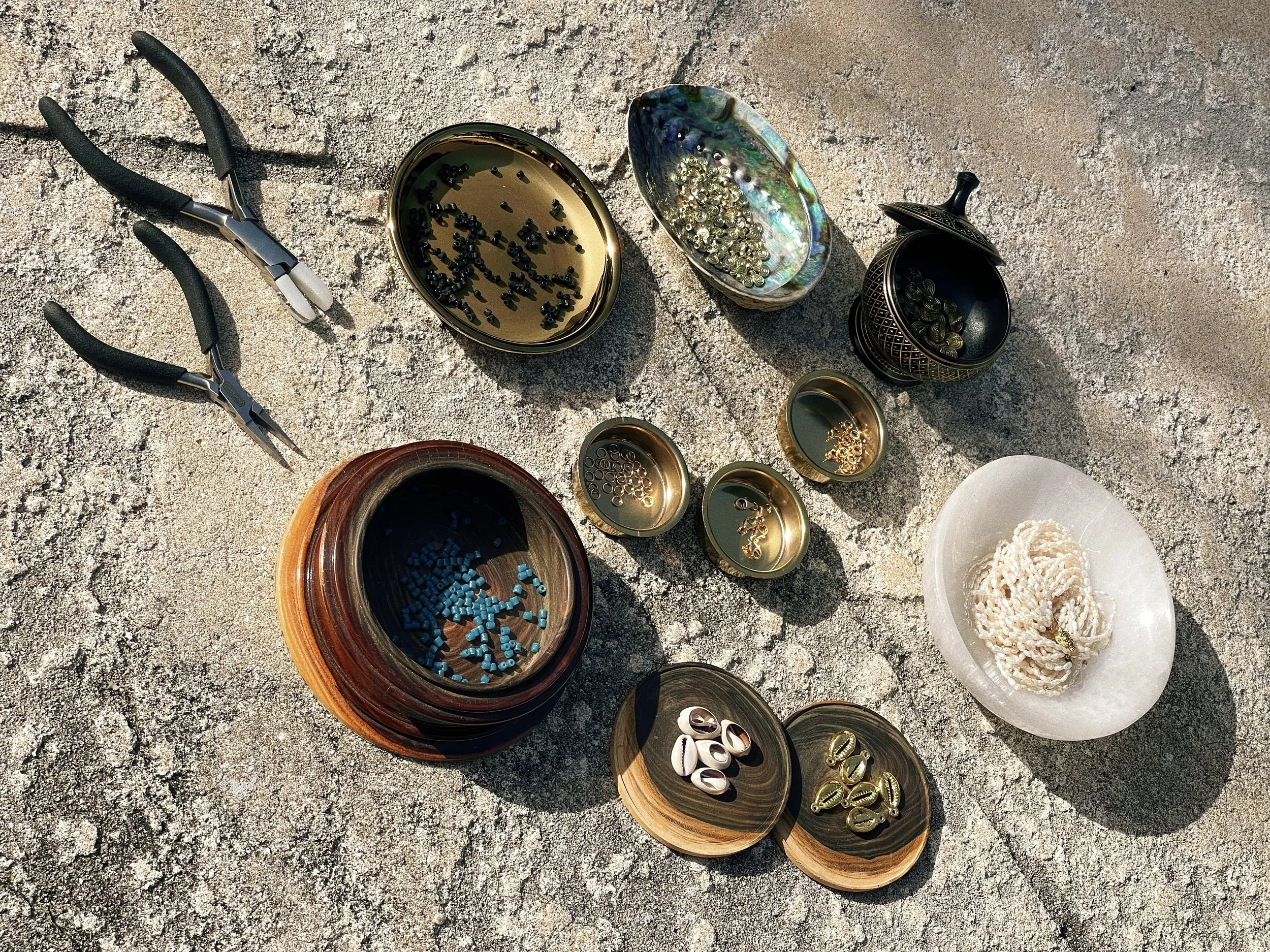 Jewelry-making materials, beads, shells, brass components, and tools arranged outdoors for the Tempered Earth creative process.