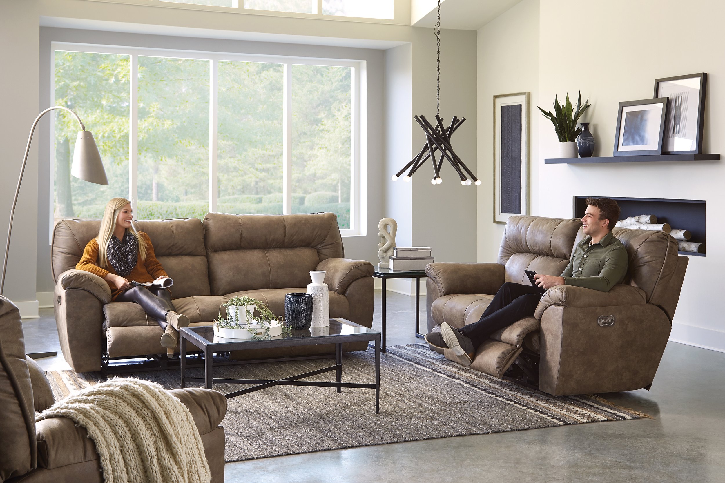 Living room with beige furniture, large window, modern chandelier, and decorative items. Two people are sitting on sofas, smiling and looking at each other.