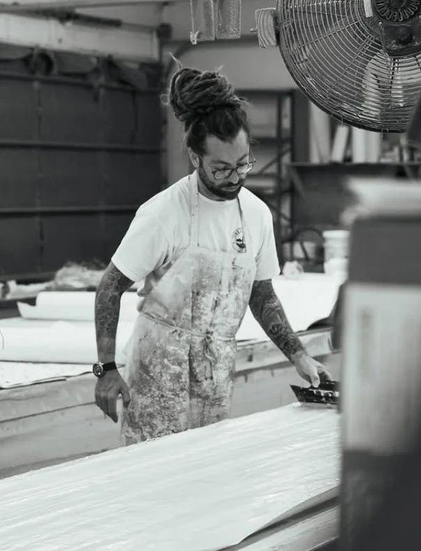A man with dreadlocks, glasses, and tattoos, wearing a white apron, working in a wallpaper factory.