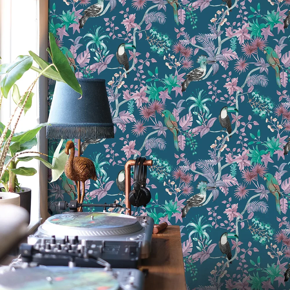 A room with a tropical-floral wallpaper featuring toucans, parrots, and hummingbirds, a DJ turntable setup, and a copper headphone stand with headphones, a blue lampshade, and potted plants nearby.