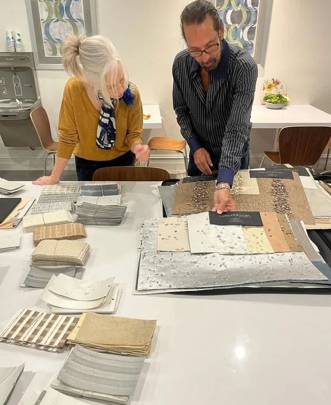 Two people, a woman with white hair and a man with glasses, are looking at fabric or wallpaper samples on a table in a room with a window and decorative wall in the background.