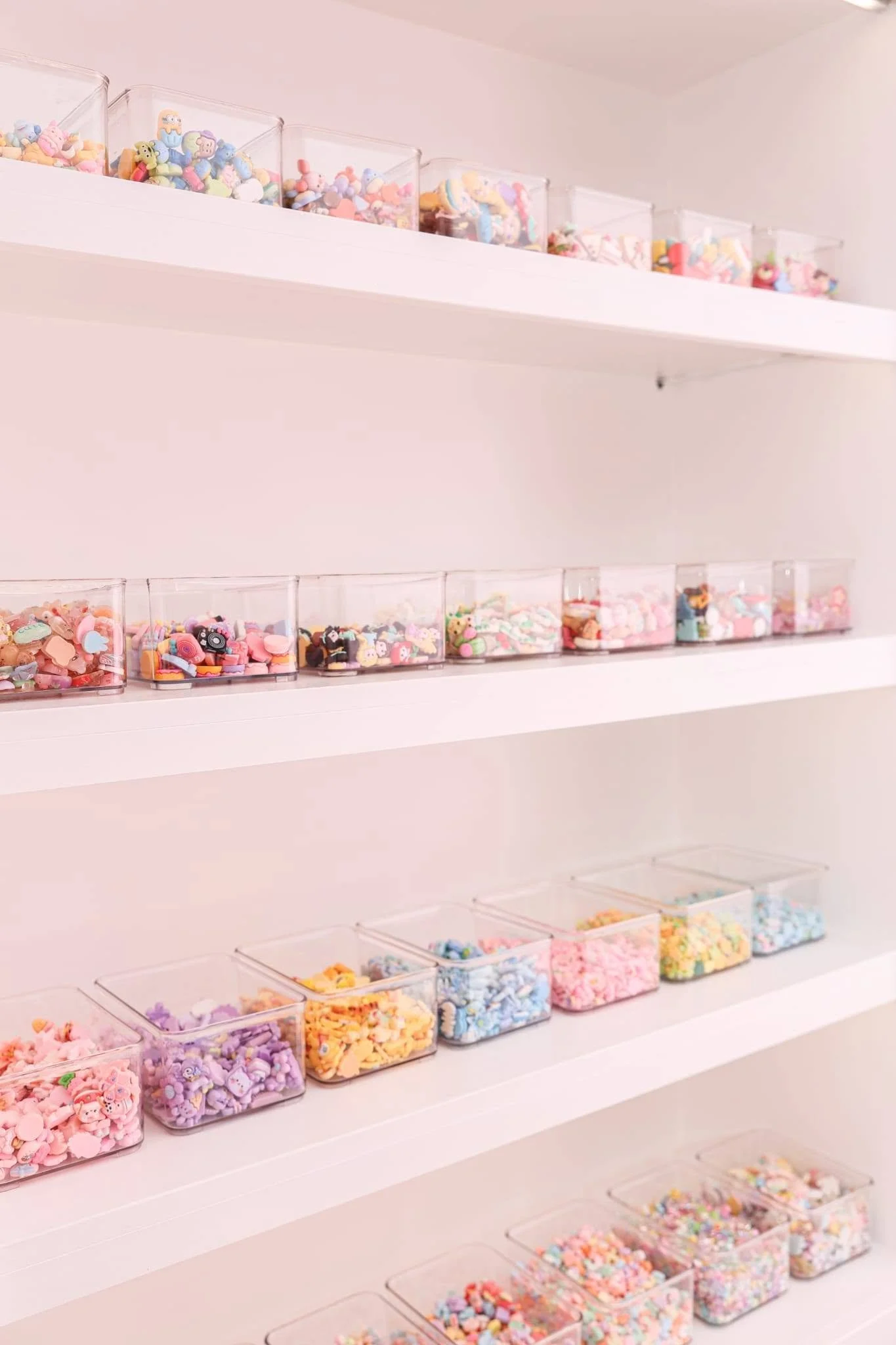 Multiple clear plastic containers on white shelves filled with colorful miniature charms and decorations, primarily in pastel shades of pink, purple, yellow, and blue.