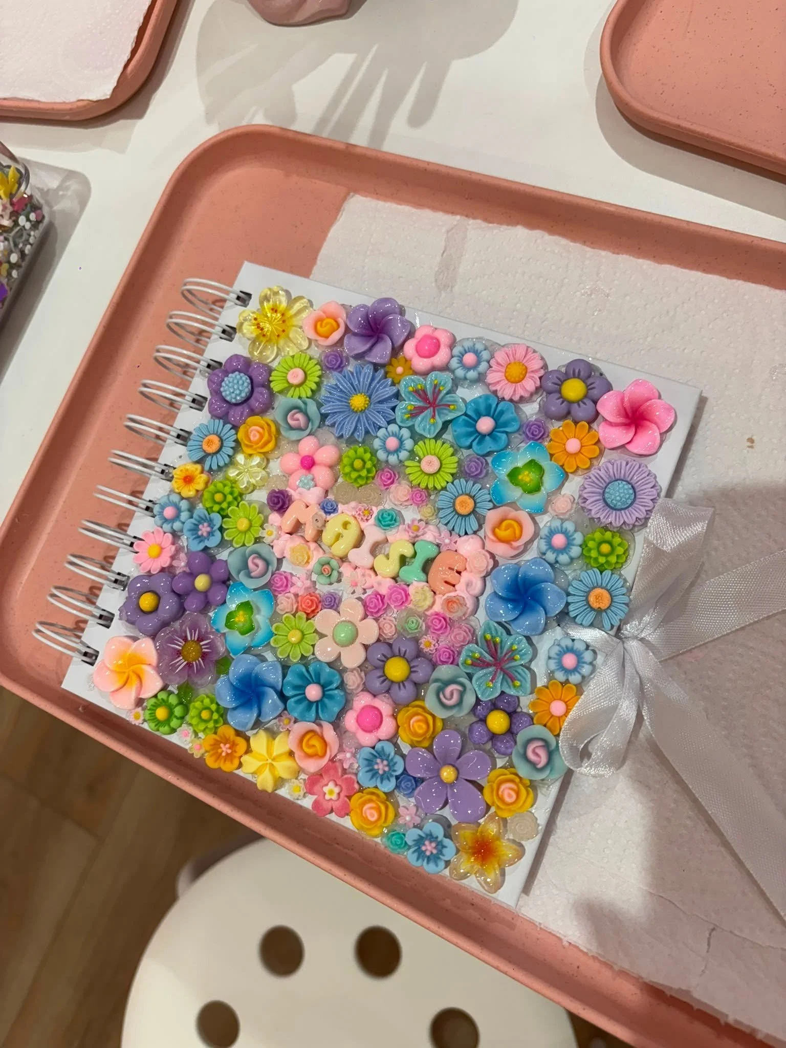 A pink tray with a white piece of paper or fabric and colorful flower-shaped objects. The objects are arranged on a white surface, with some overlapping and others spaced apart, and are in various bright colors including purple, blue, pink, yellow, o