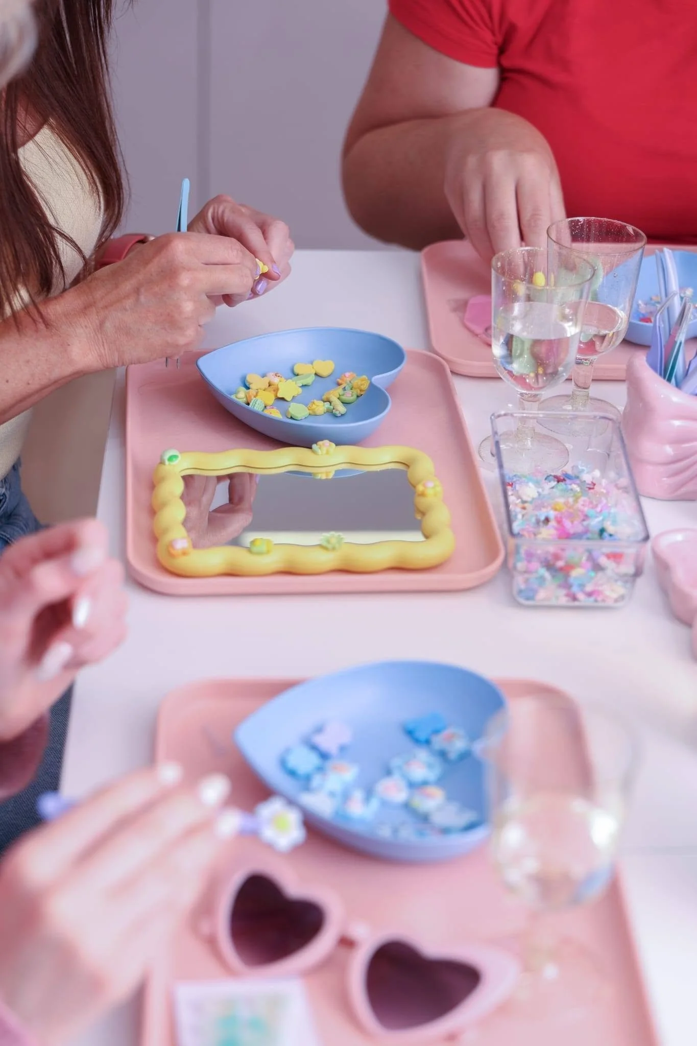 Close-up of two people decorating heart-shaped and other colorful sugar cookies with small decorative candies at a table with pink and blue trays and cups with water.