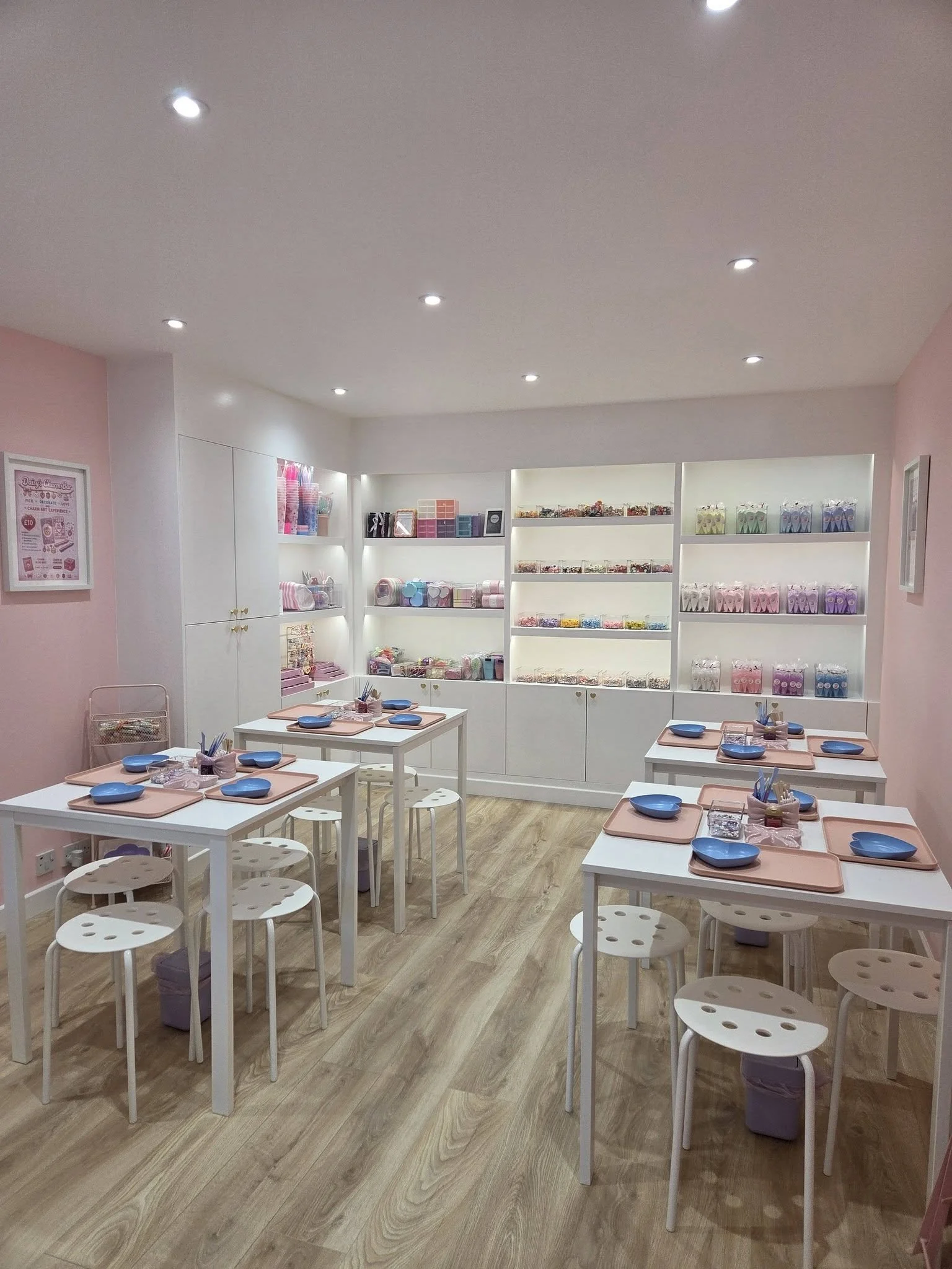 A brightly lit, pastel-colored classroom with white tables, blue bowls, stools, and shelves filled with craft supplies and decorative items.