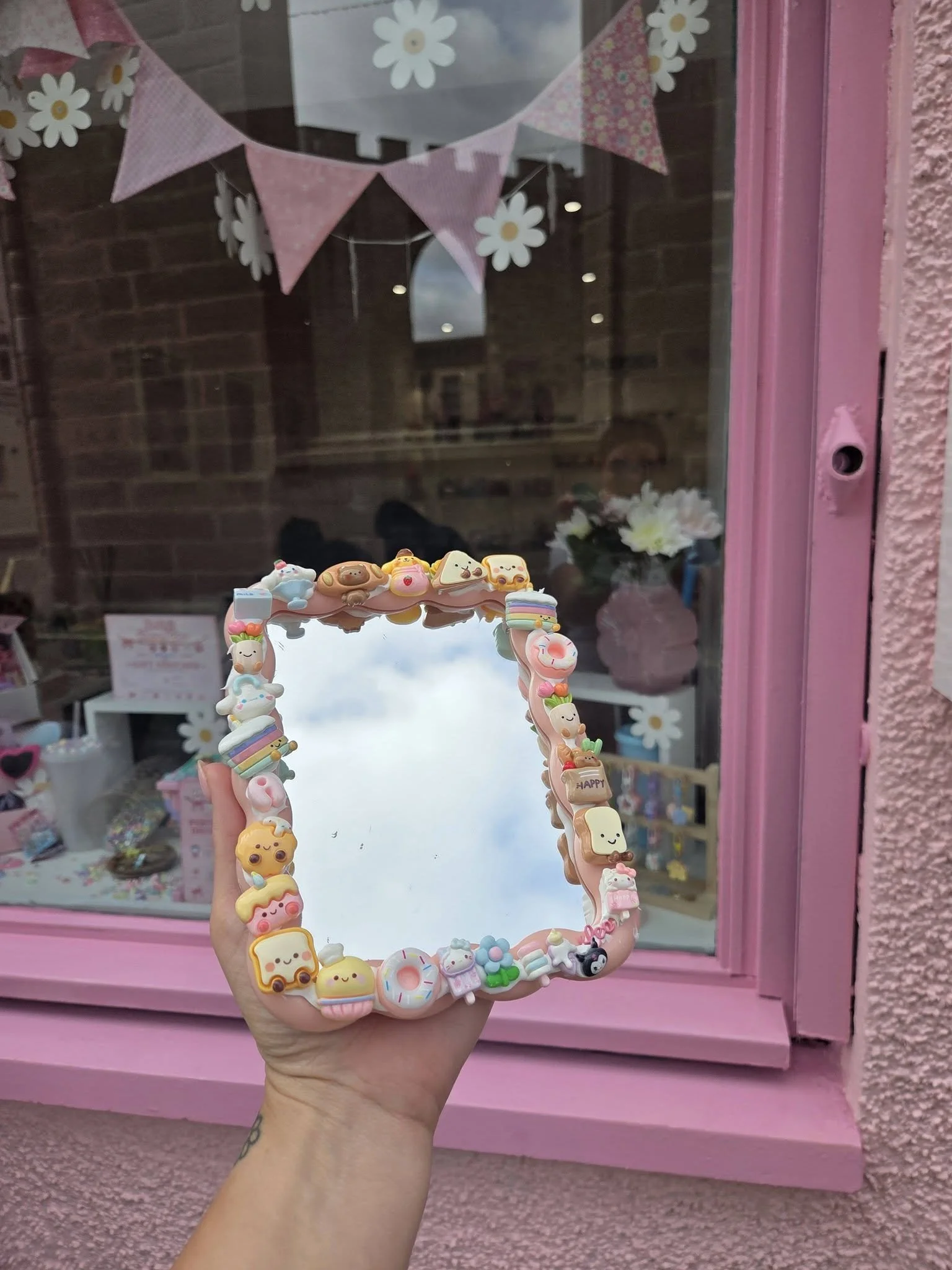 Hand holding a pink mirror with kawaii-themed charms, with a pink window frame, and a store with pink decor and flowers in the background.