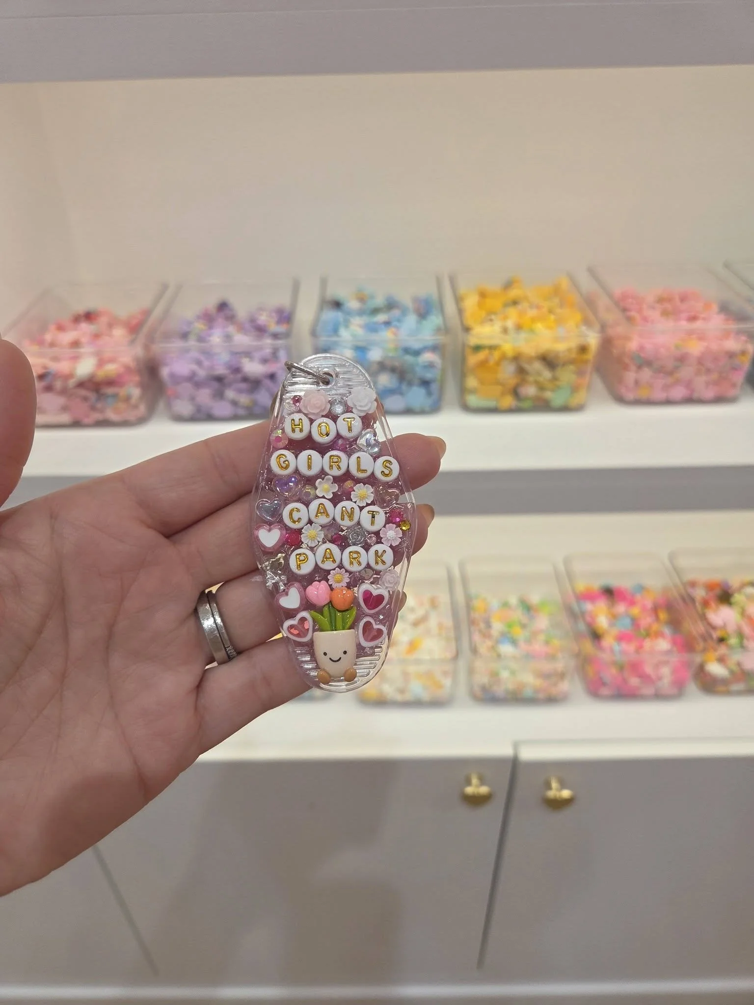 Hand holding a clear plastic keychain with the phrase 'HOT GIRLS CAN'T PARK' spelled out in gold letters decorated with small pink hearts, flowers, and a smiling flower pot at the bottom. In the background, there are small transparent containers fill