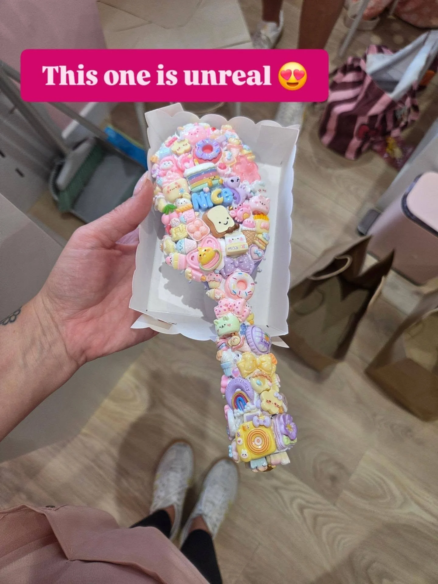 A long candy hairbrsuh made of pastel-colored charms, including rainbows, clouds, unicorns, and other cute figures, hung over a hand holding a container of decorated charms. The background shows a room with chairs, tables, and some people.