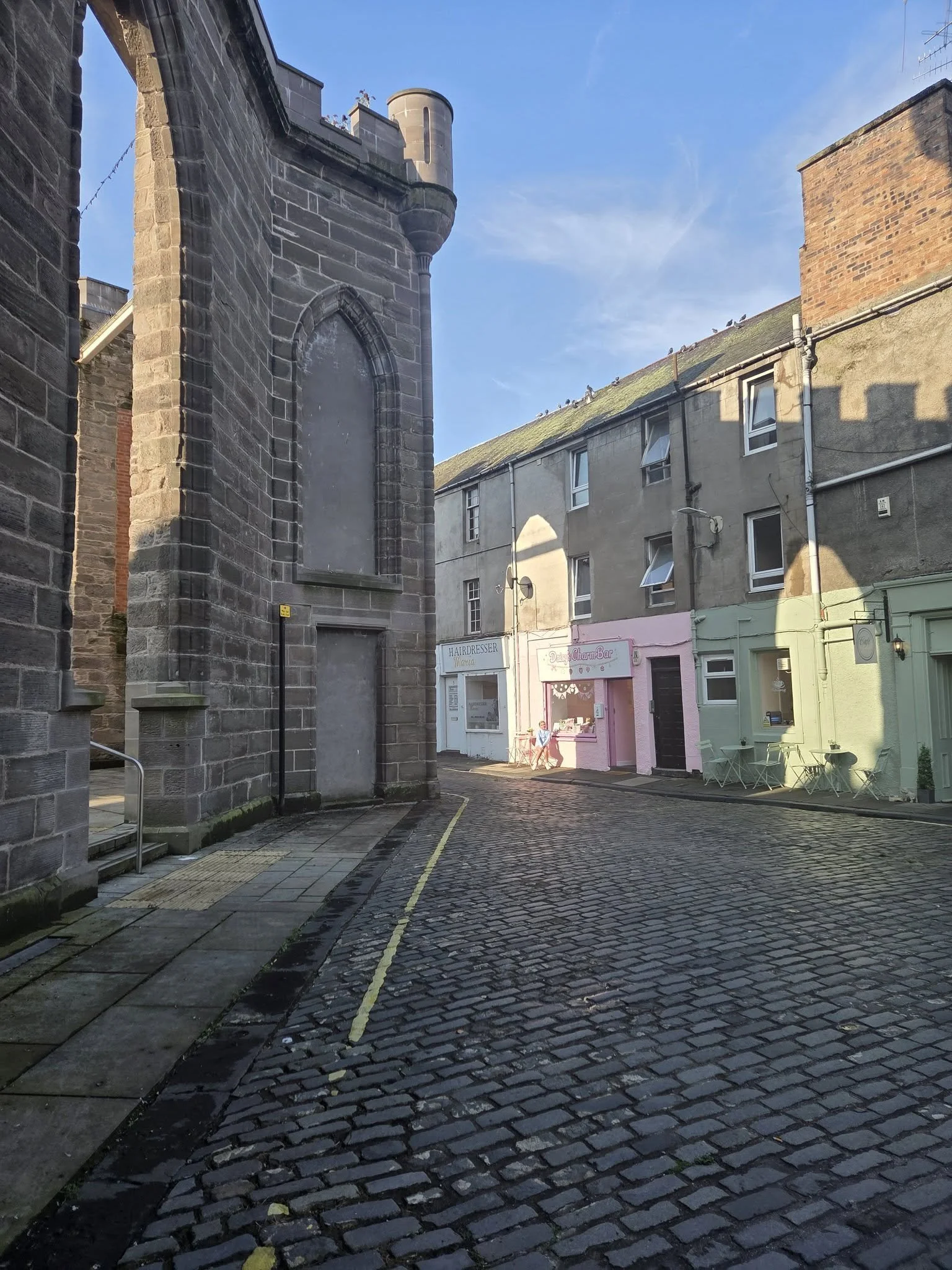 A narrow cobblestone street in a small town, with a stone church or historic building on the left, pastel-colored shops and cafes on the right, and a clear blue sky above.
