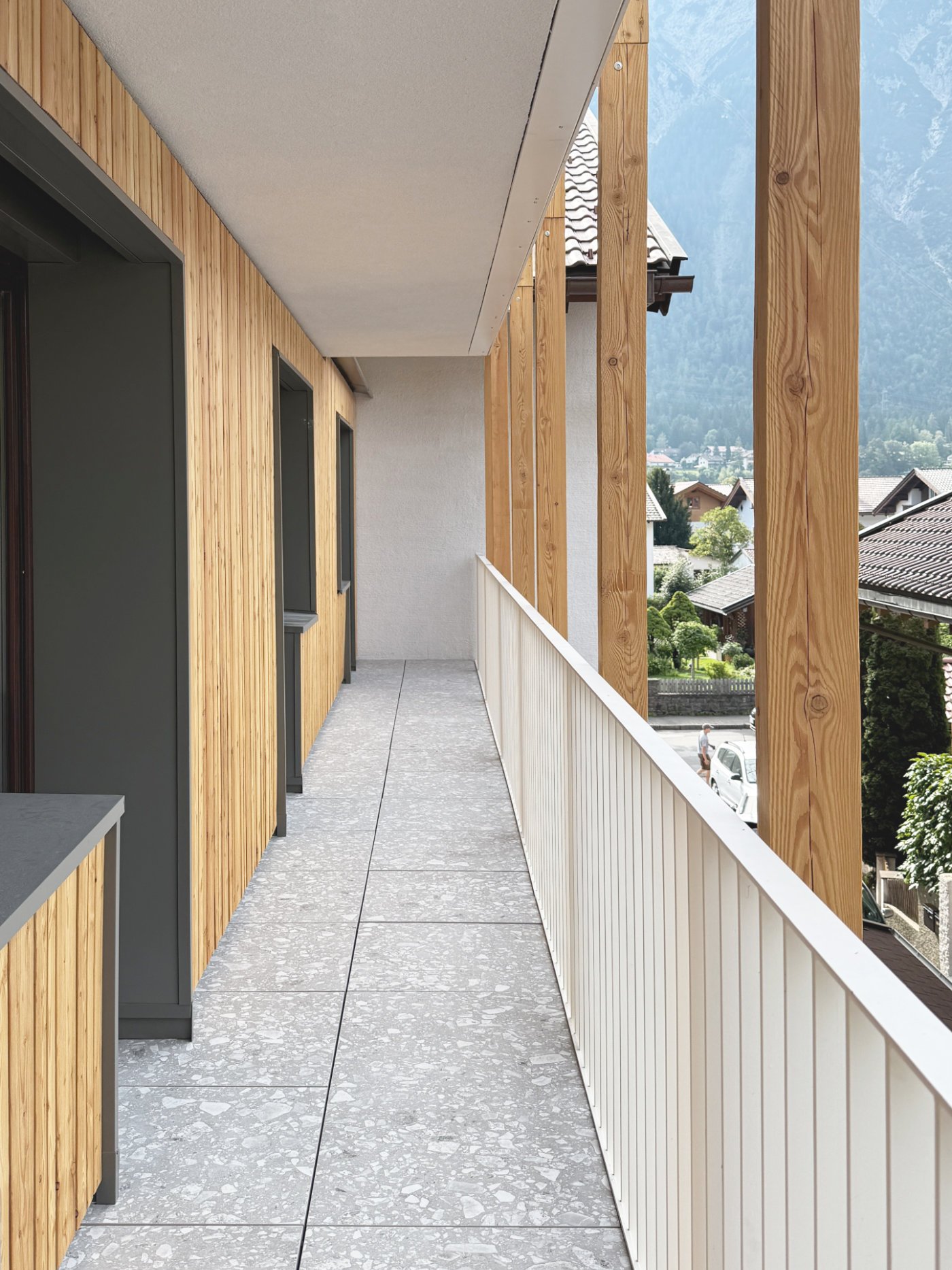 Durchgehender Balkon mit Holzfassade und dunkelgrauen Fensterlaibungen mit Ceppo di gré Fliesenboden im Mitarbeiterhaus J in Mittenwald, geplant von d'ns Studio
