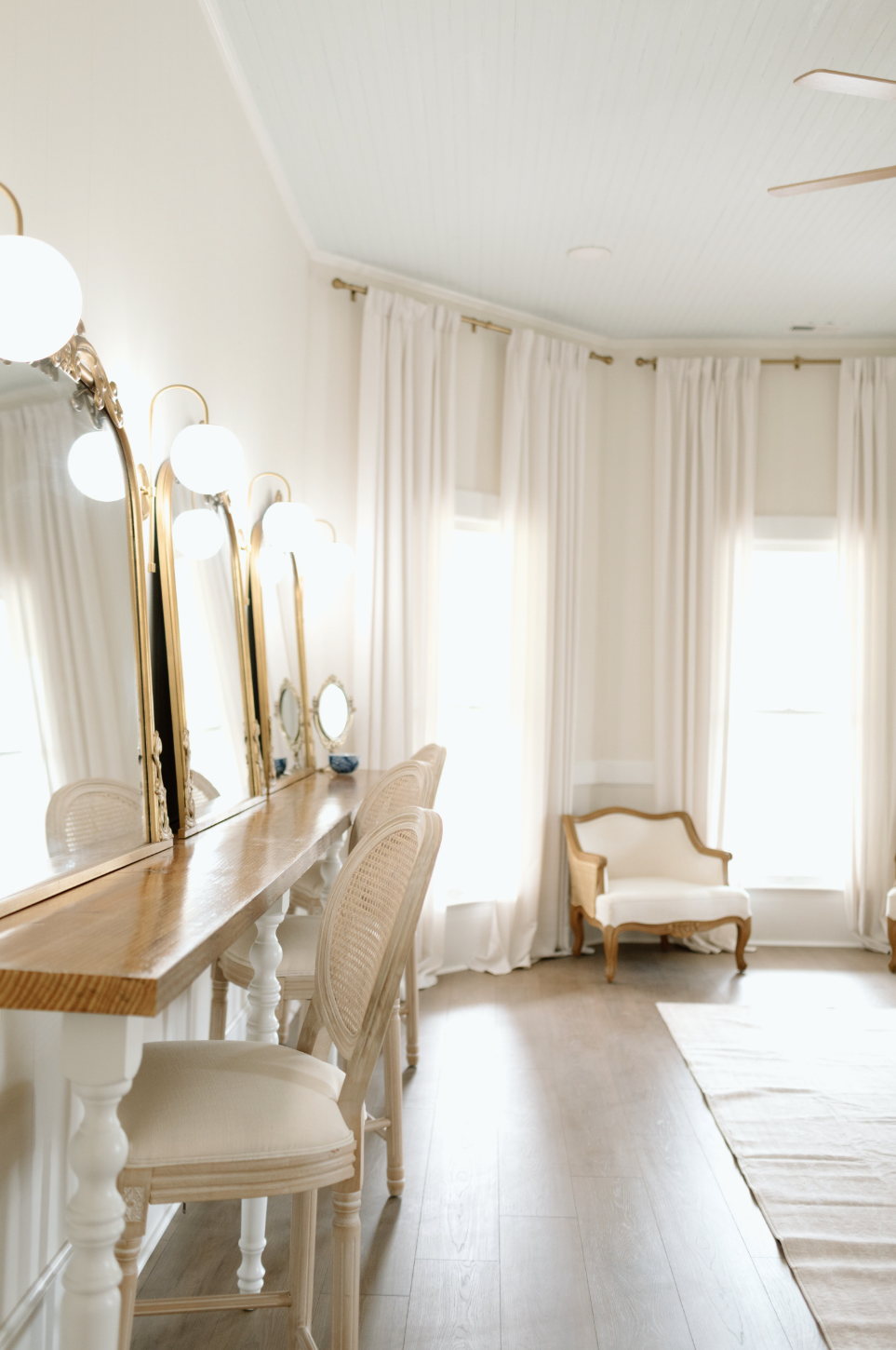 A bright dressing room with a wooden vanity, three oval mirrors with gold frames, and three white globe lights. Two upholstered chairs are in front of the vanity and a vintage-style loveseat is by the window, with white curtains and a light-colored r