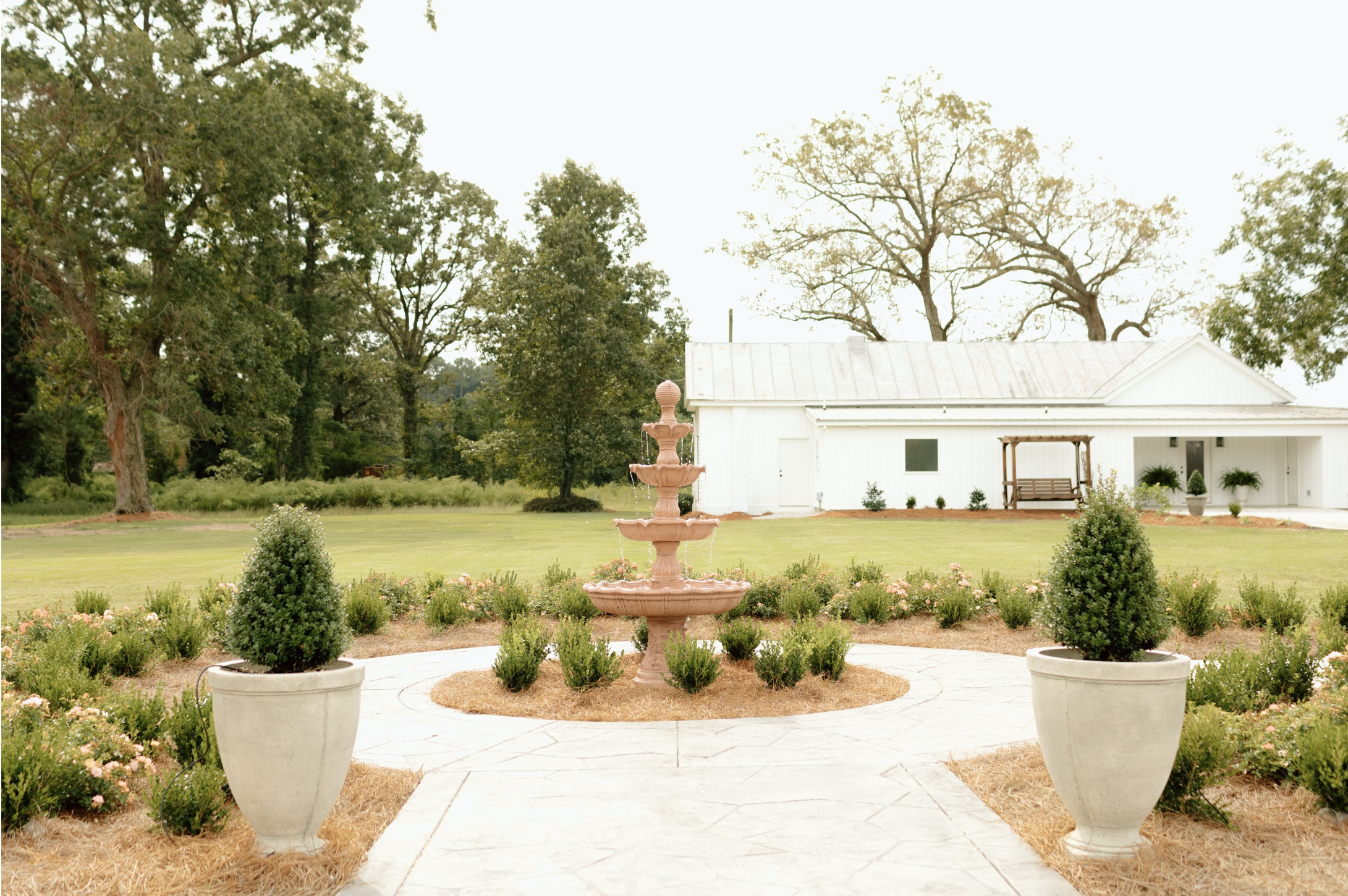 A garden with two large white pots containing small bushes, a three-tier pink fountain, a manicured lawn, a white building in the background, and several tall trees under a cloudy sky.