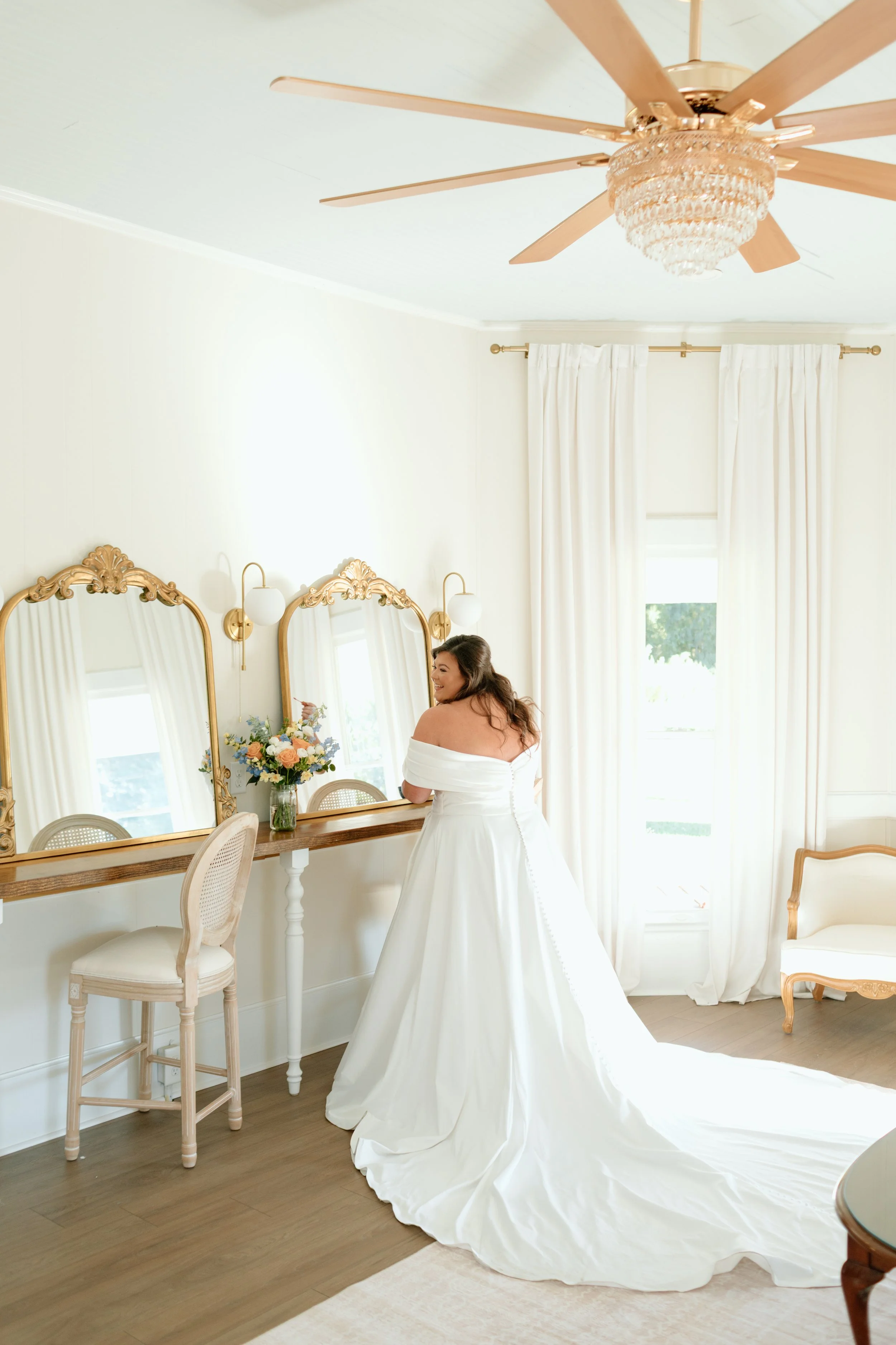 A woman in a white wedding dress standing in a bright room with elegant decor, looking in a mirror, surrounded by flowers and vintage furniture.