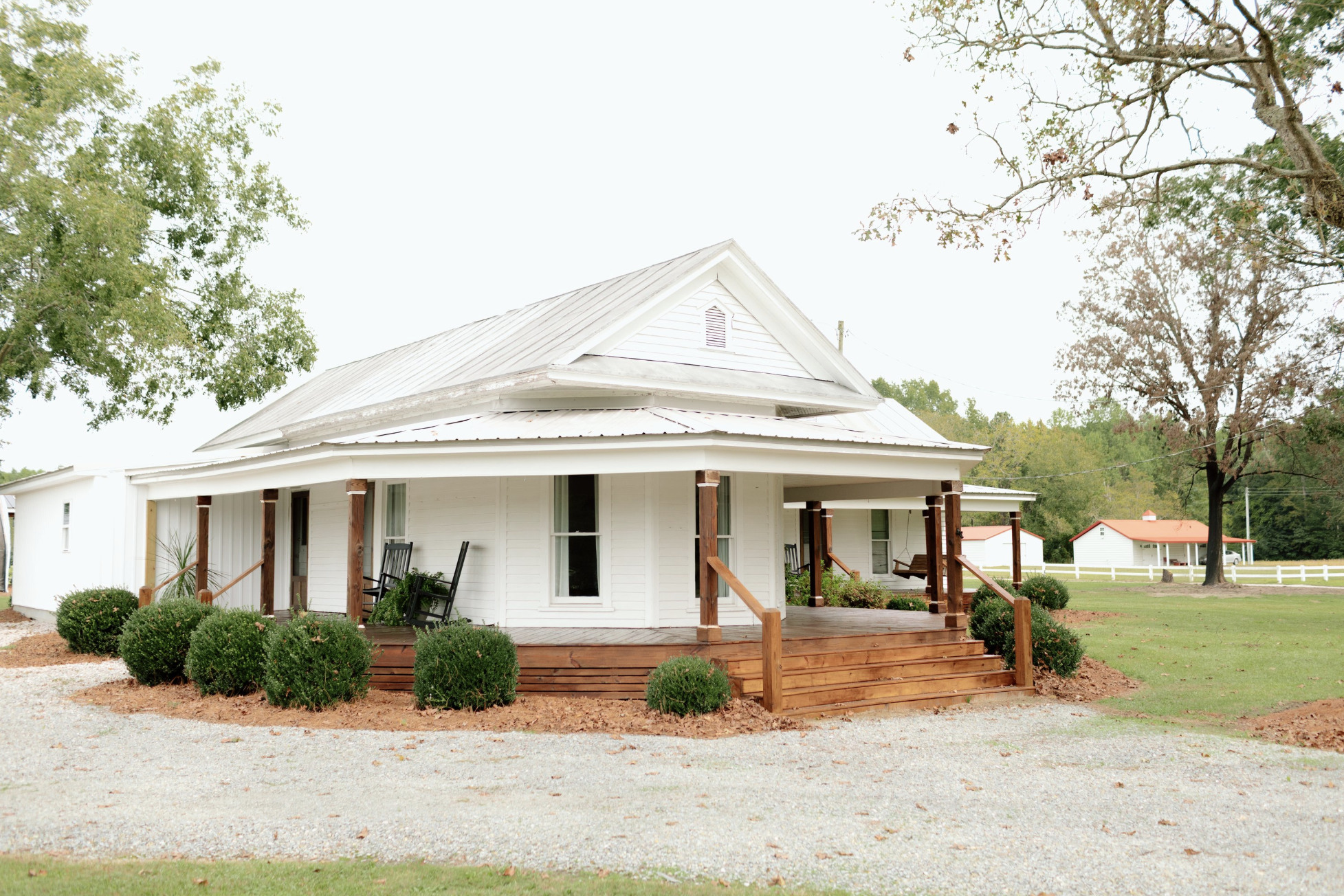 A white house with a metal roof, front porch with rocking chairs, green bushes, and trees, under a cloudy sky.