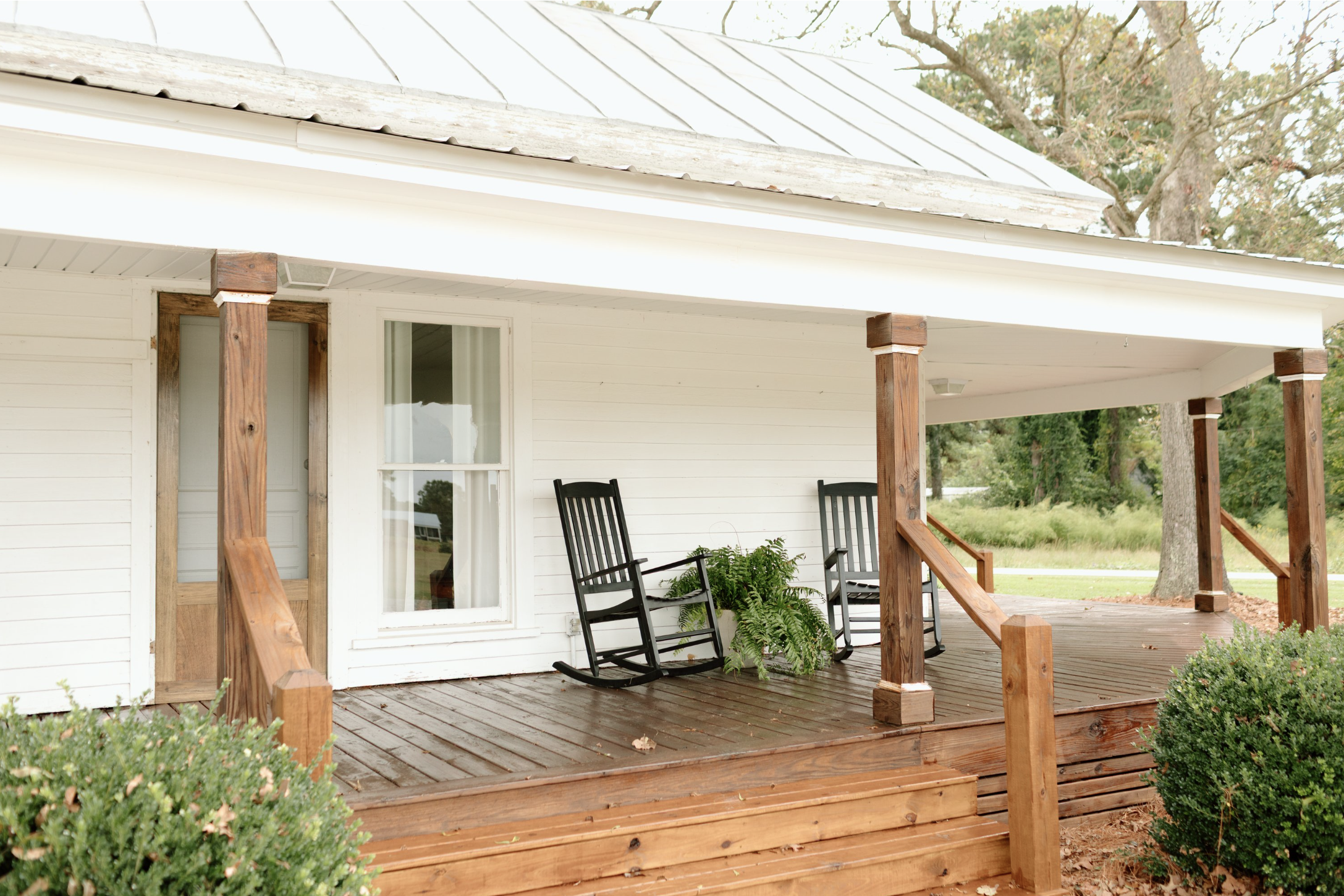 A porch with wooden steps and railing, two black rocking chairs, a large window on a white house, and trees in the background.
