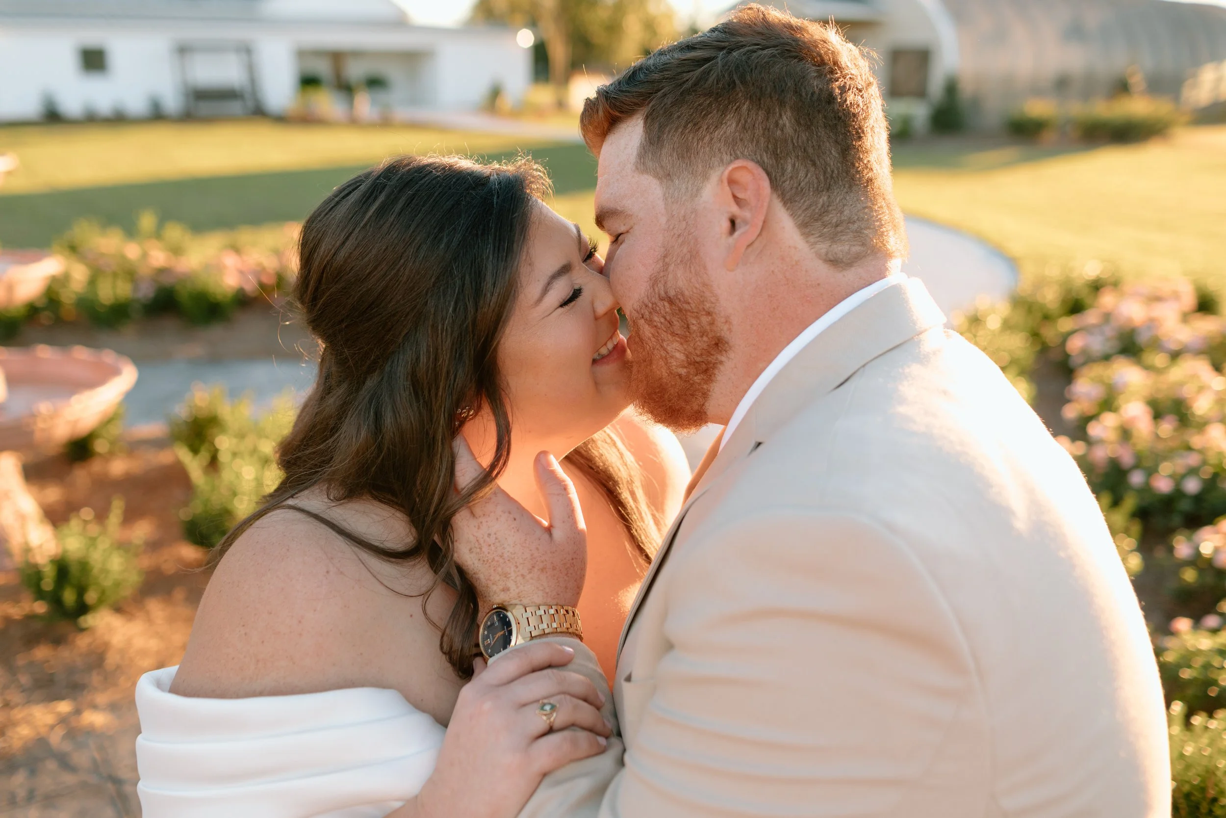 A couple is sharing an intimate moment outdoors, touching foreheads and smiling with their eyes closed during golden hour in a garden setting.
