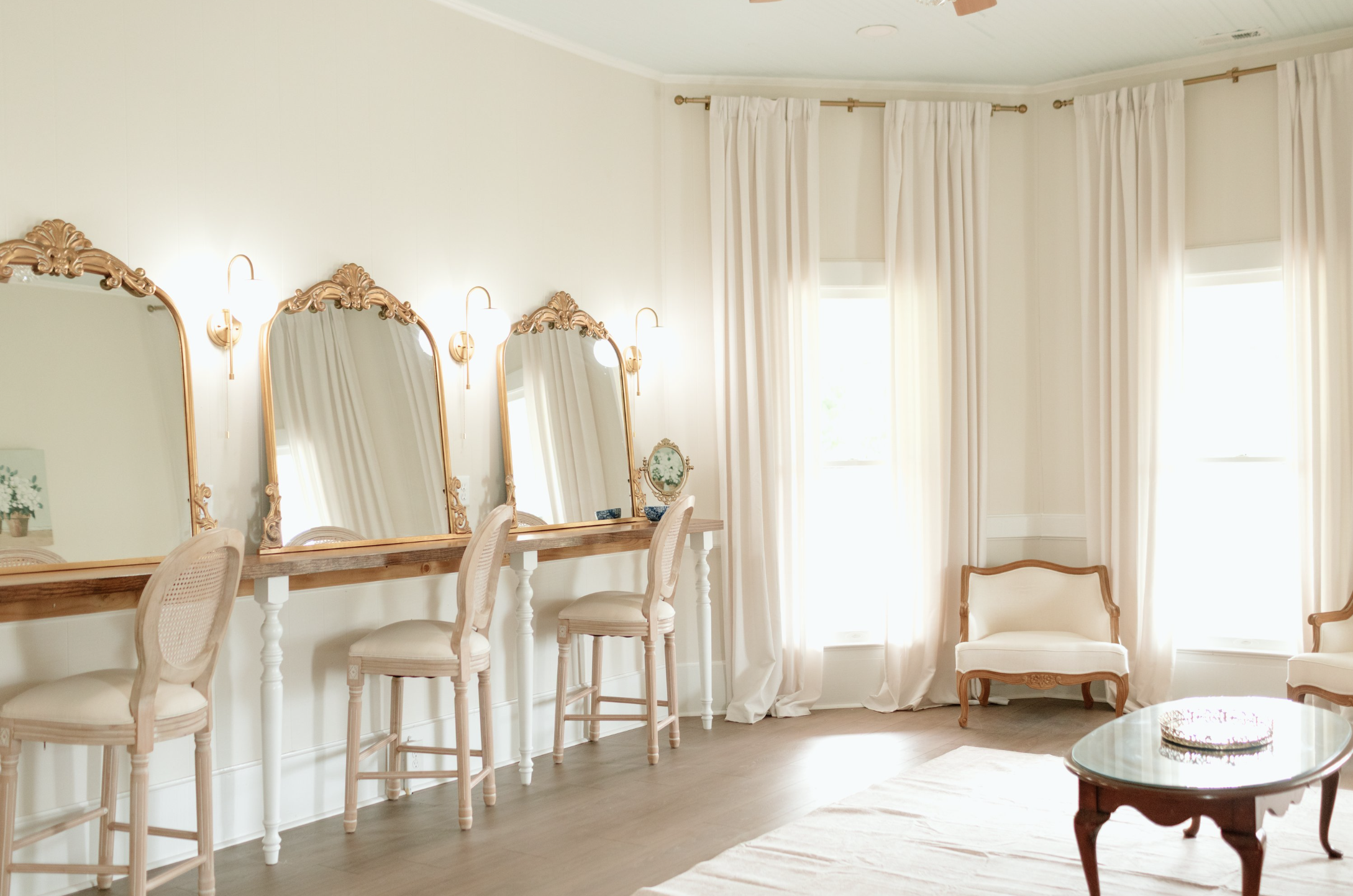 A bright living room with white walls, large windows with white curtains, and a wooden bar-height table with wrapped chairs. There are three decorative mirrors with ornate gold frames hanging on the wall above the table. Small lamps are mounted next 