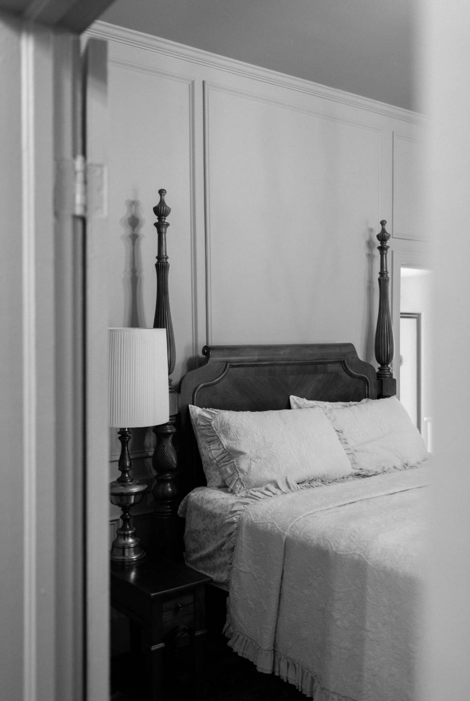 A black-and-white image of a bedroom with a wooden four-poster bed, nightstand, and lamp, viewed through an open door.
