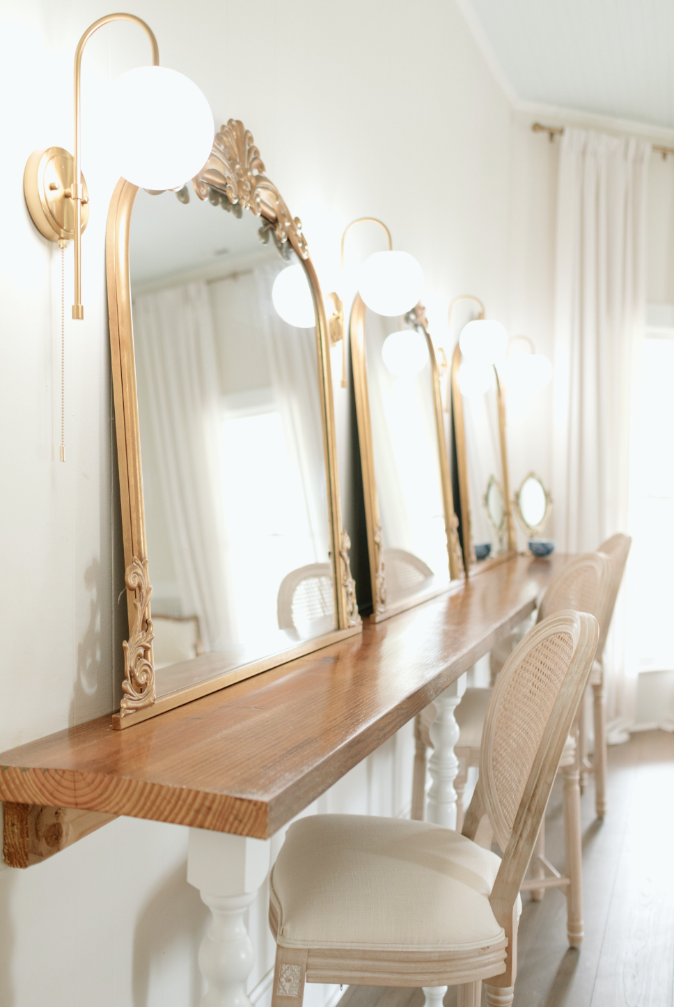 A wooden vanity table with four ornate mirrors and two wall-mounted light fixtures with globe bulbs in a room with white curtains and light-colored furniture.