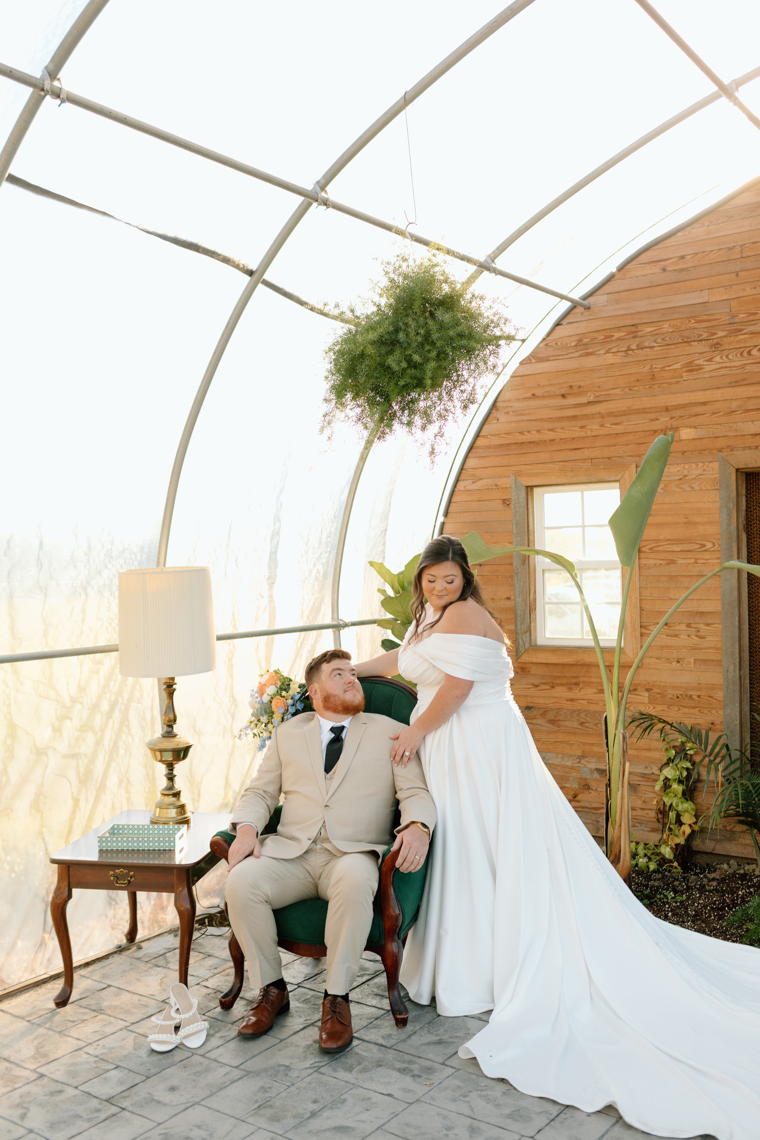 A woman in a white wedding dress is standing next to a man in a beige suit sitting in a green armchair in a greenhouse. There are large green plants, a wooden wall, and sunlight in the background. A pair of white high-heeled shoes is on the floor.