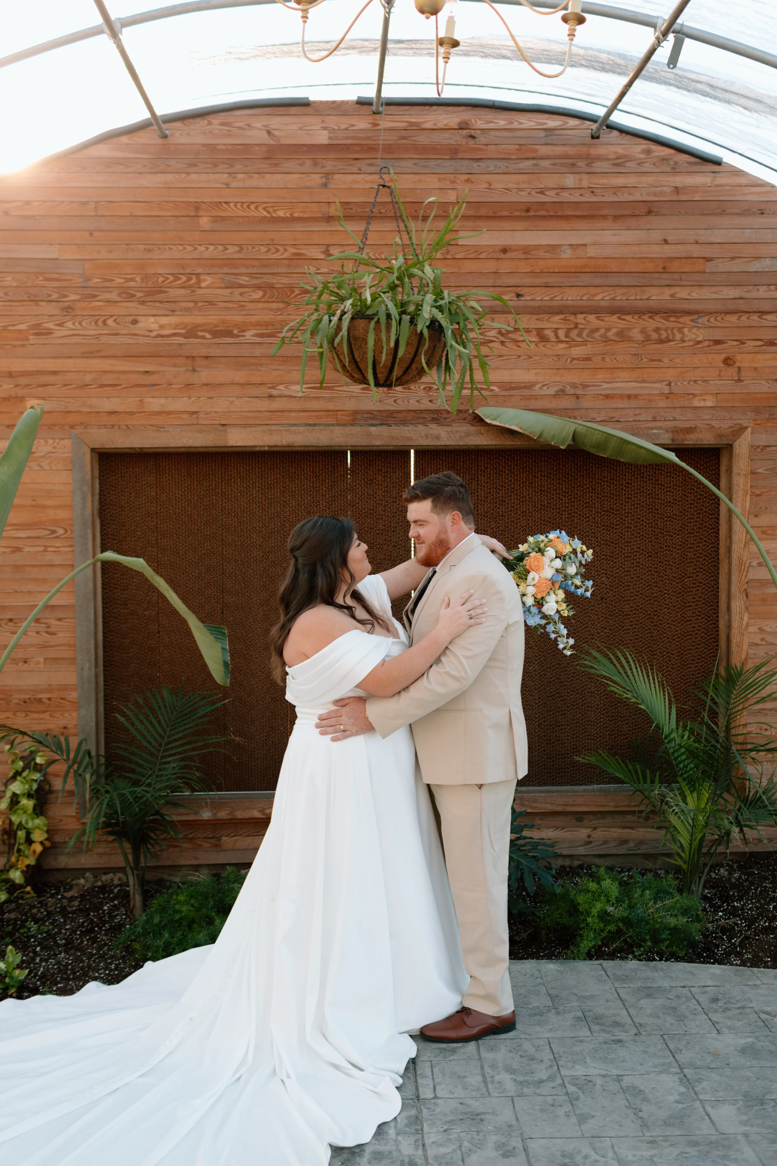 A bride and groom share a dance outdoors, surrounded by tropical plants, with a wooden wall and hanging plant overhead.
