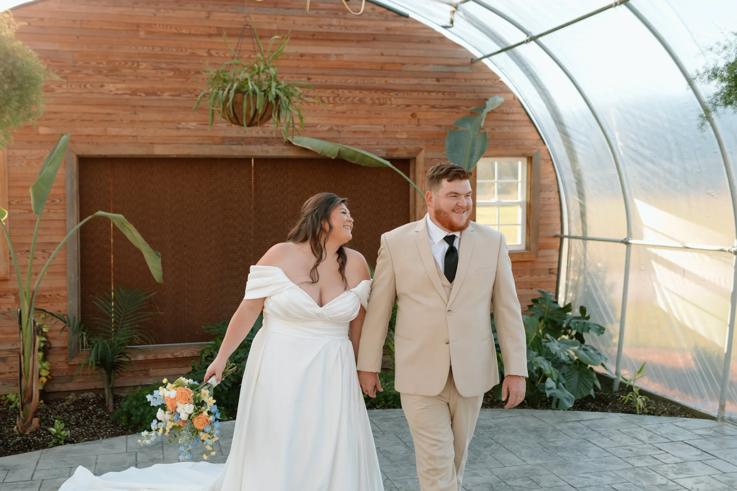 A bride and groom walking together in a greenhouse, smiling and holding hands. The bride wears a white off-shoulder wedding dress, and the groom wears a beige suit with a black tie. The bride holds a bouquet of yellow, orange, and blue flowers, and t
