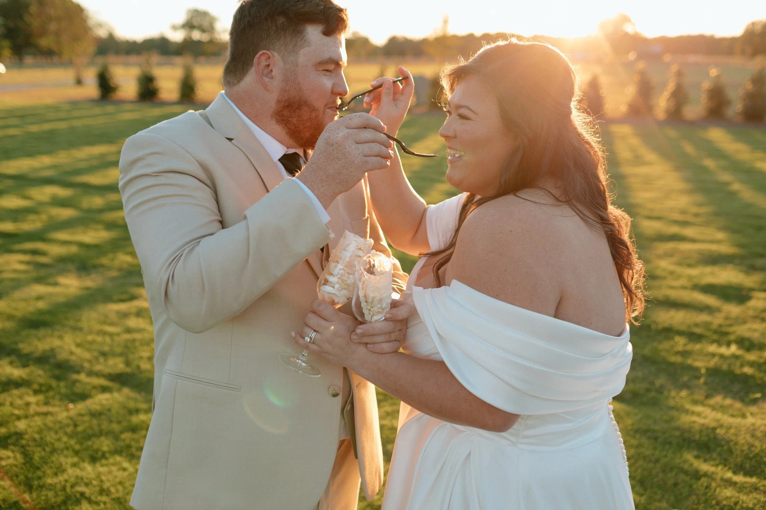 A newlywed couple outdoors at sunset, with the groom feeding the bride a piece of cake. They are smiling and embracing, with a grassy field and trees in the background.