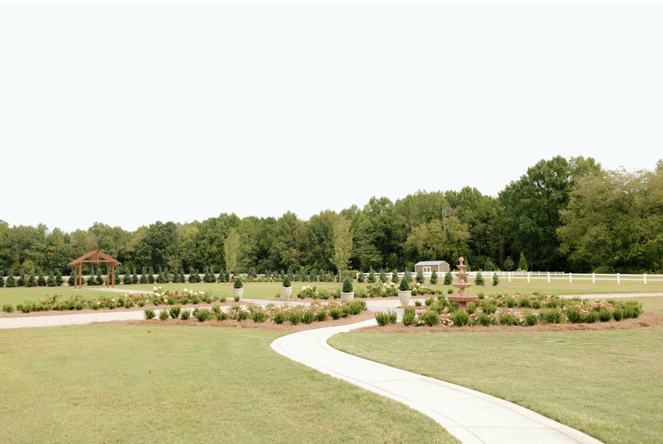 A landscaped garden with a curved concrete pathway, flowerbeds, potted plants, a fountain, a wooden gazebo, trees, and a white fence under a clear sky.