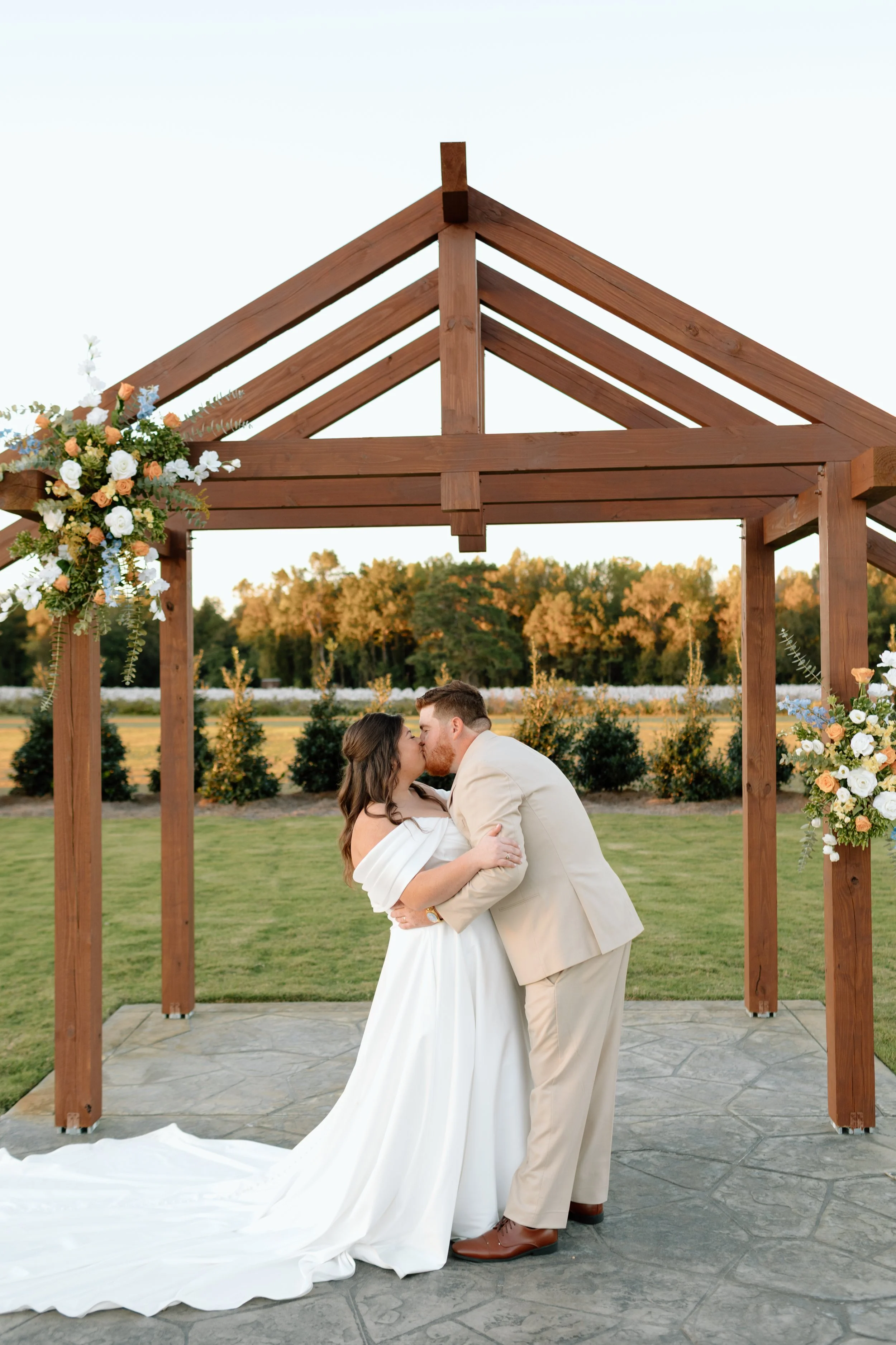 A bride and groom kissing beneath a wooden arch decorated with flowers during an outdoor wedding ceremony.
