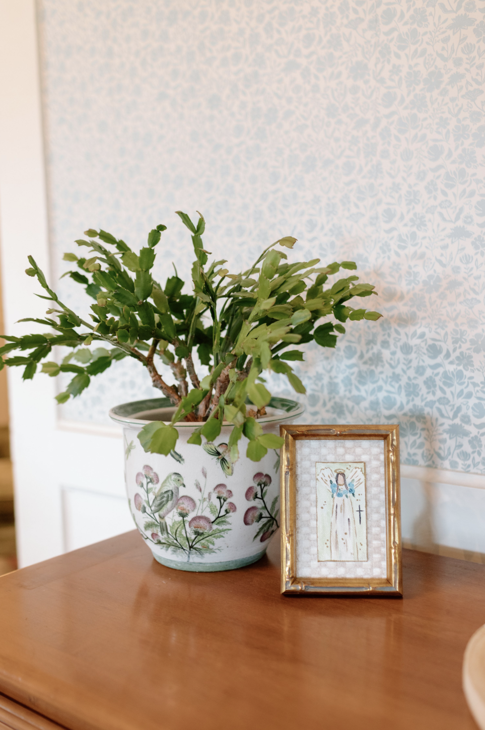 A potted plant with green leaves on a polished wooden table, next to a framed religious cross and image, in a room with light blue floral wallpaper.