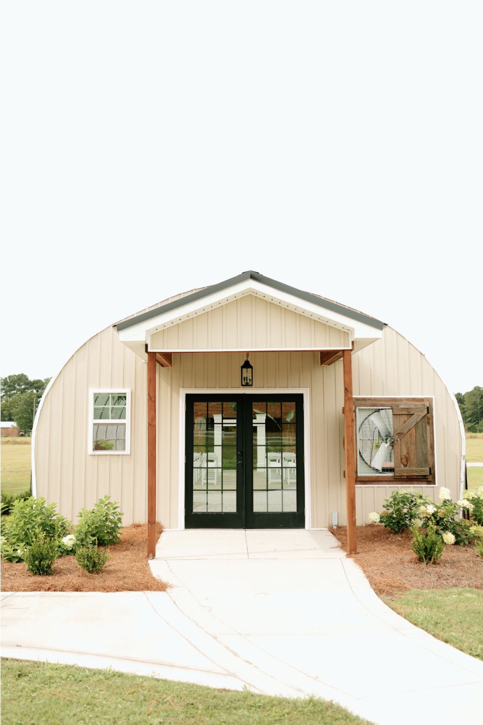 A small, cream-colored, barn-style building with a black door, small windows, and a satellite dish on the right side. It has a porch with wooden posts and is surrounded by grass and small bushes.