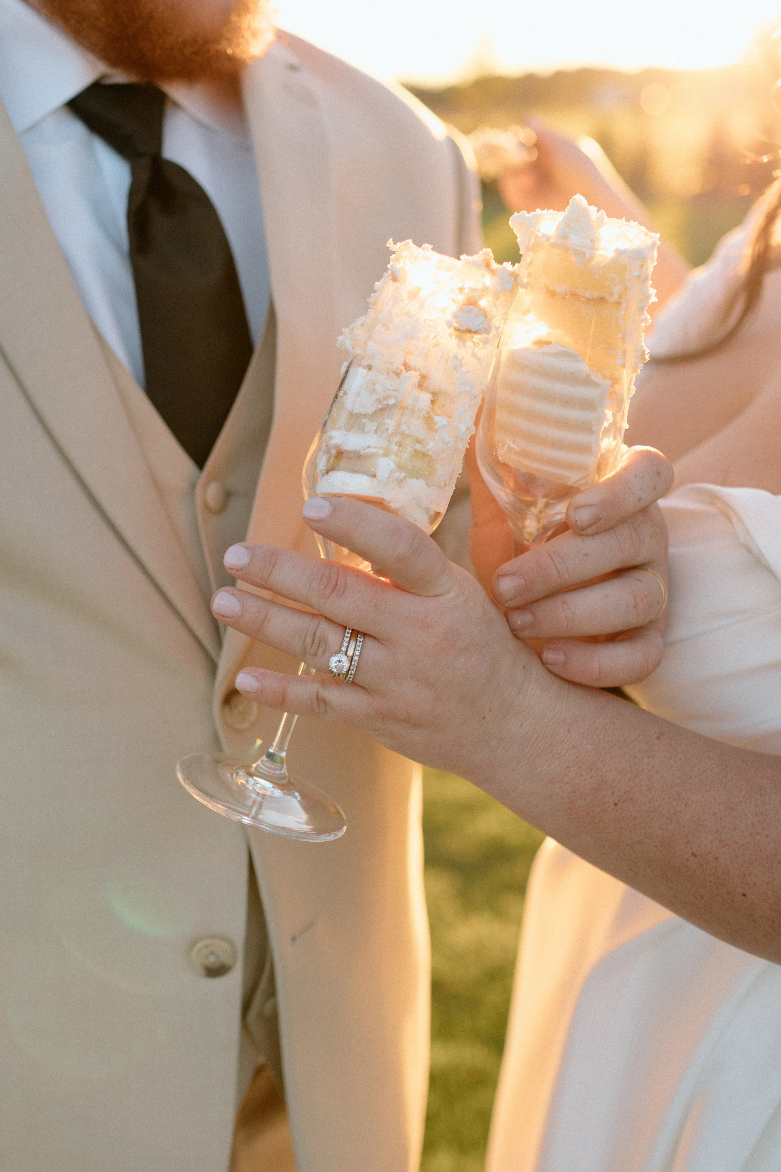 A close-up of a person's hand holding two champagne glasses filled with a layered dessert, with a man dressed in a beige suit and black tie in the background during a sunset outdoor event.