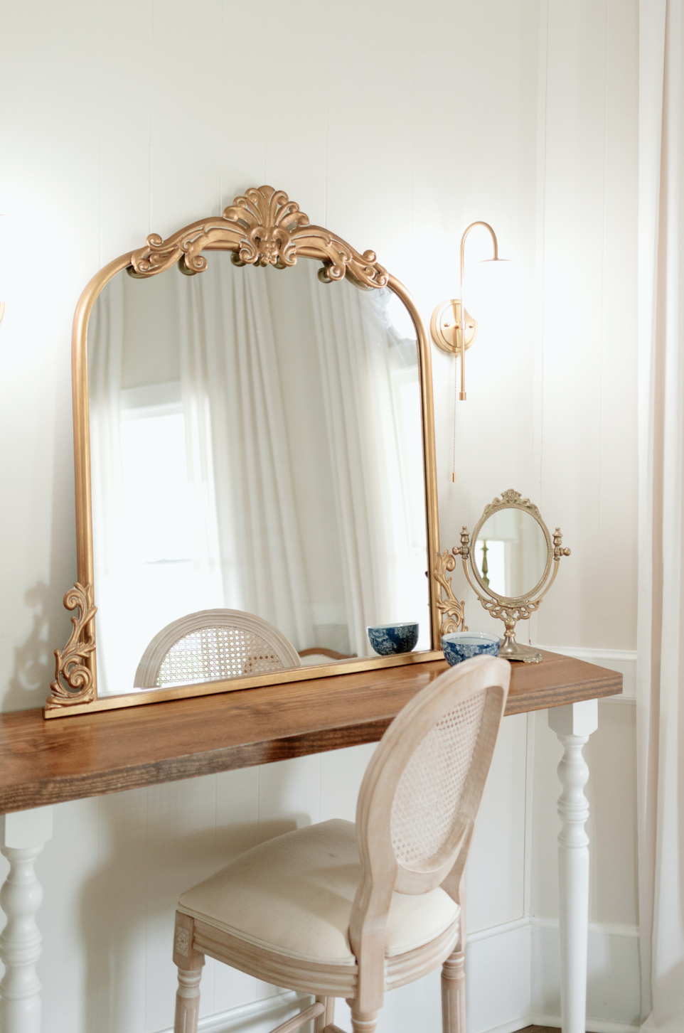 A vintage wooden vanity table with a large decorative mirror, a small oval mirror, two blue and white bowls, and a chair with a woven cane back is positioned against a cream-colored wall with a sconce and curtains reflected in the mirror.