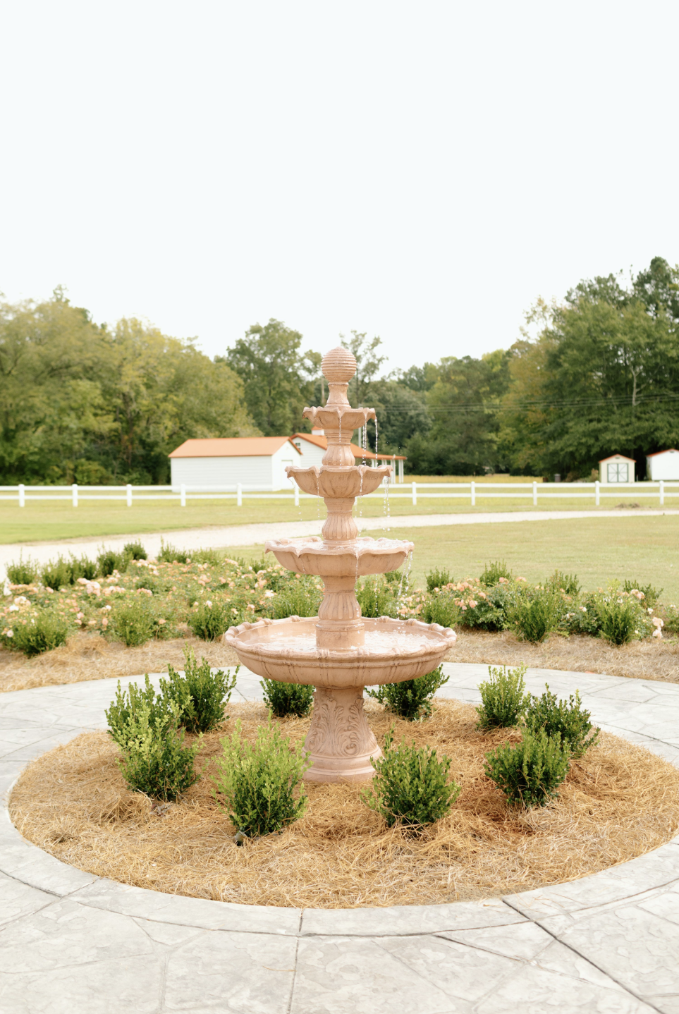 A pink multi-tiered fountain in a garden with bushes and flowers, surrounded by a stone pathway and enclosed by a white picket fence with a barn and trees in the background.
