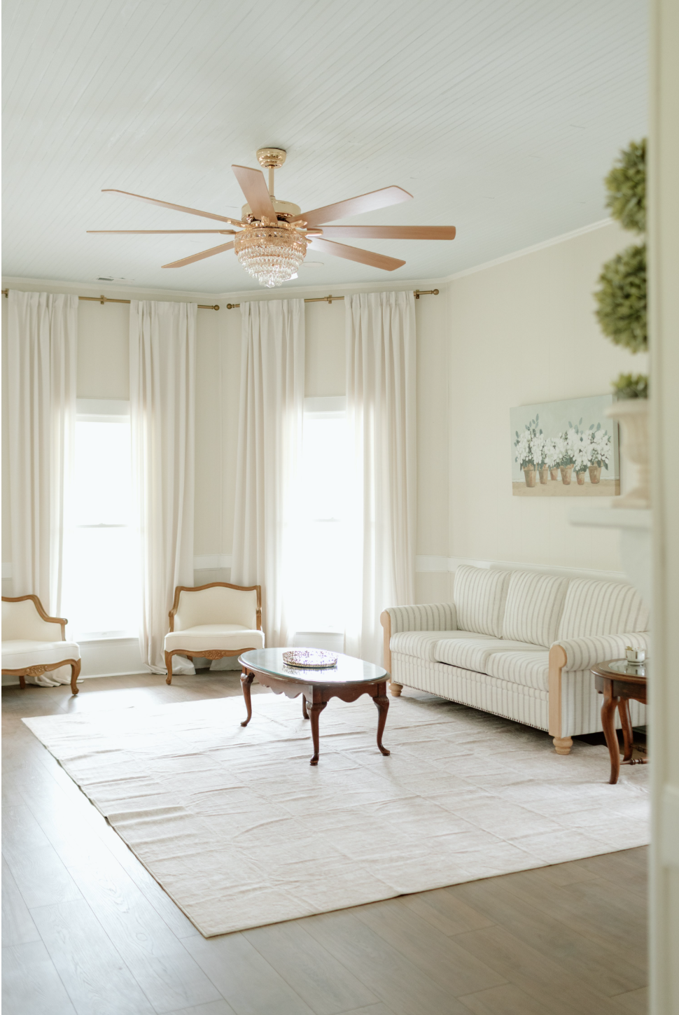 Bright living room with three large windows, cream-colored curtains, a white striped sofa, two matching armchairs, a wooden coffee table, a small round side table, and a ceiling fan with a crystal light fixture.