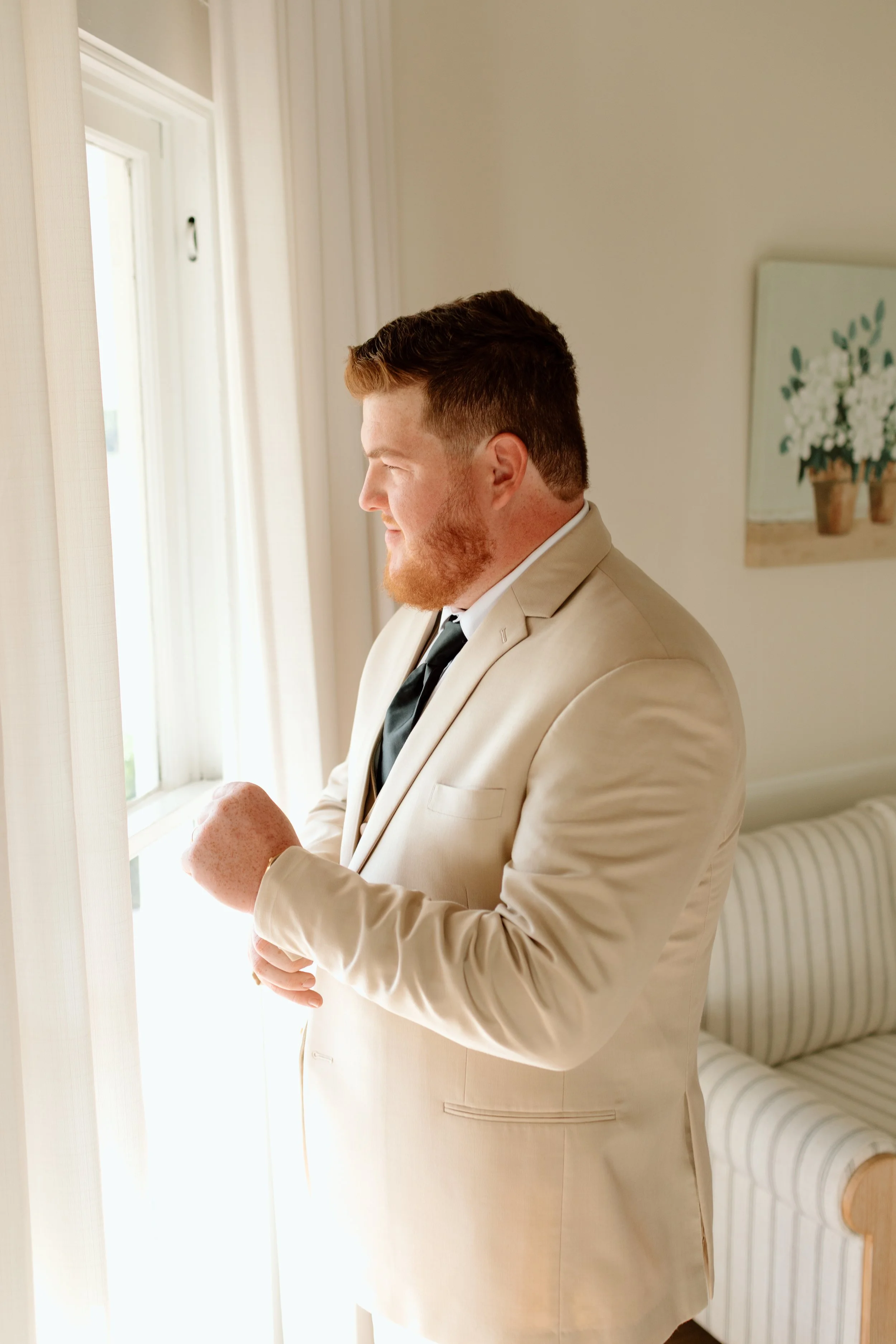 A man with red hair and beard wearing a cream suit, white shirt, and black tie, standing by a window, looking outside, with hands clasped. Behind him is a striped sofa and a canvas with white flowers on the wall.