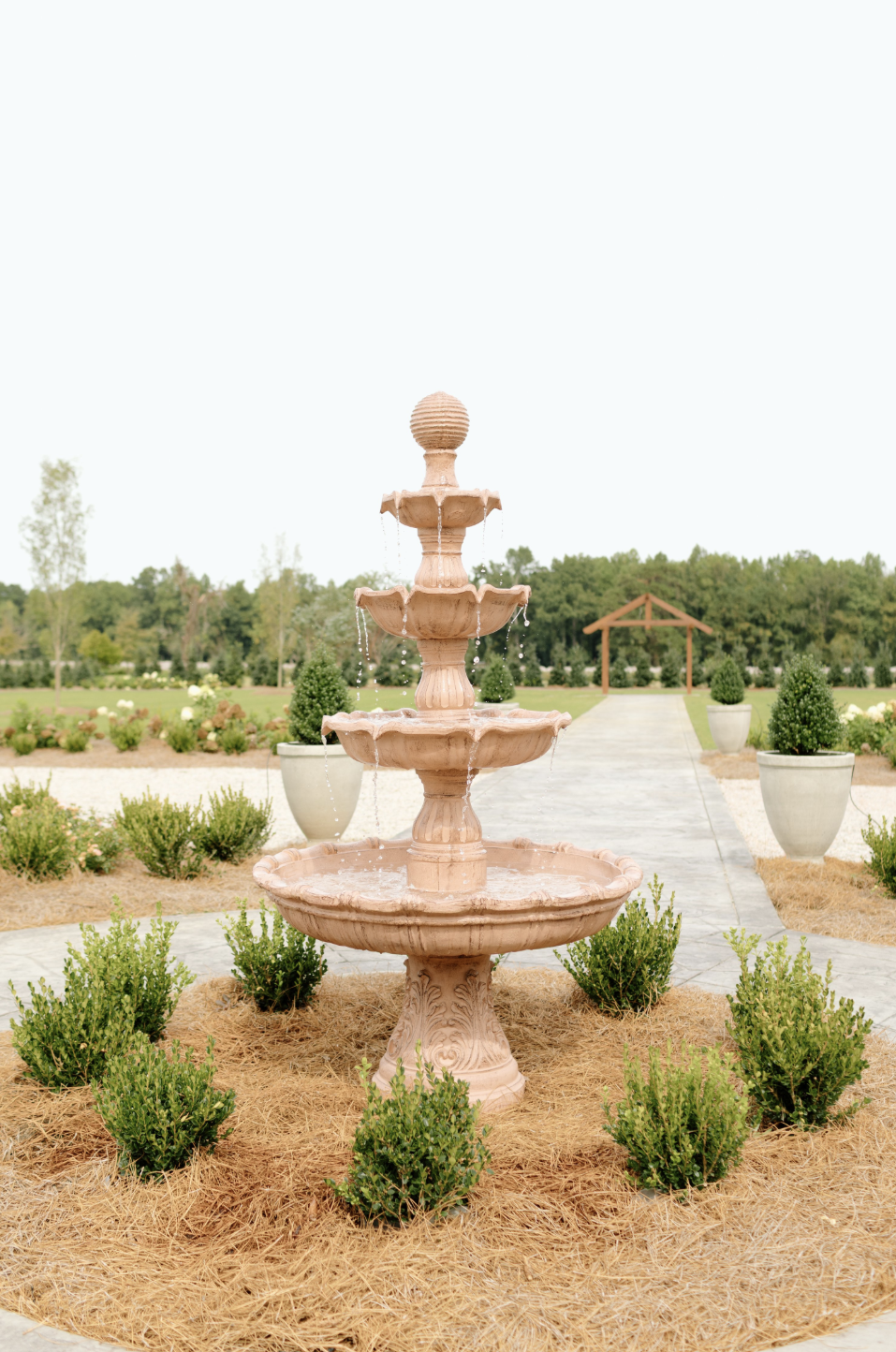 A pink multi-tiered outdoor fountain in a landscaped garden with potted plants and a pathway leading to a wooden arbor.