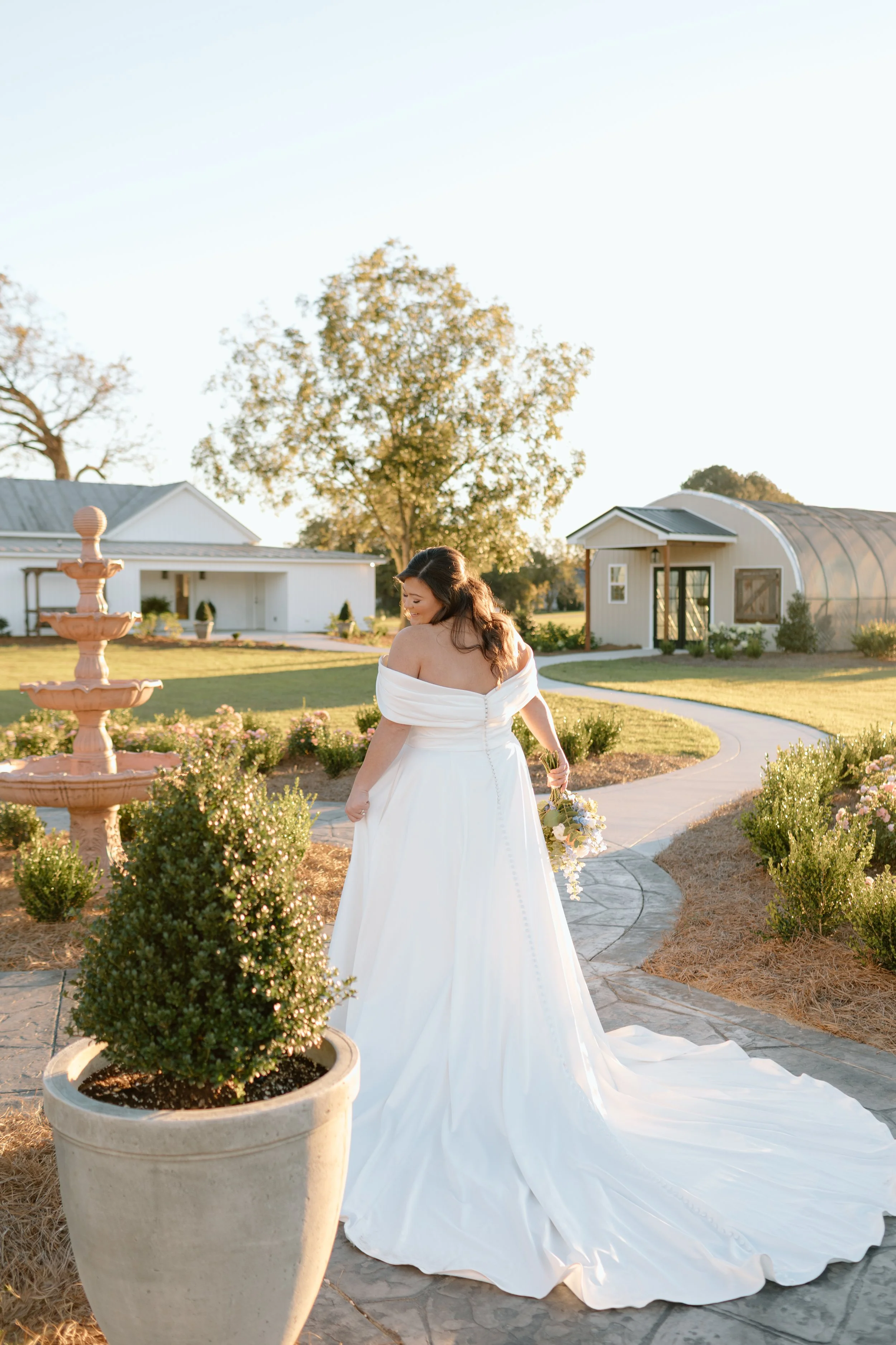 A woman in a wedding dress holding a bouquet of flowers, standing outdoors near a potted plant with a garden and buildings in the background during sunset.
