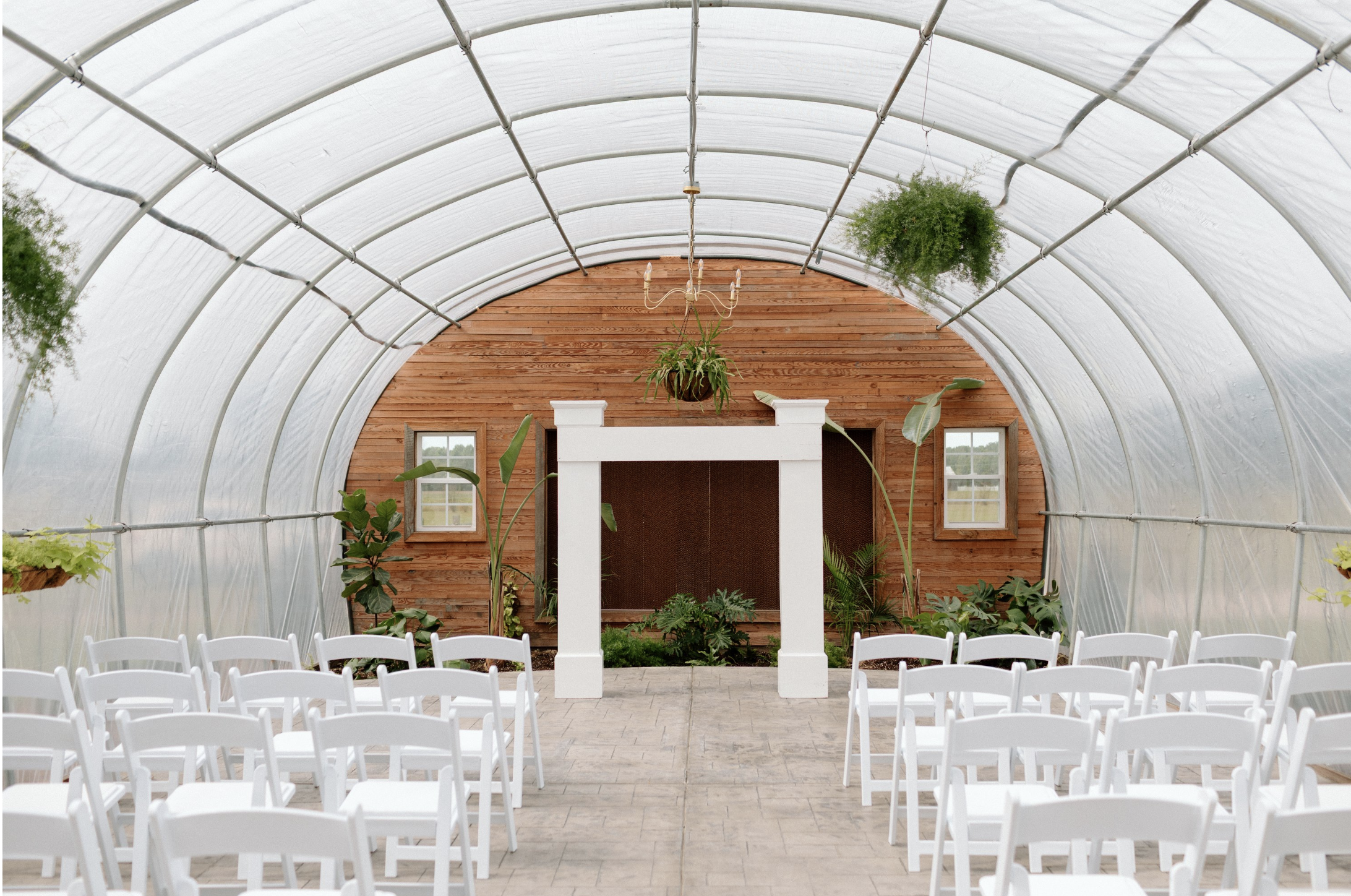 Indoor greenhouse with a curved roof, white chairs arranged in rows, potted plants, hanging greenery, wooden wall, window, and chandelier chandelier.