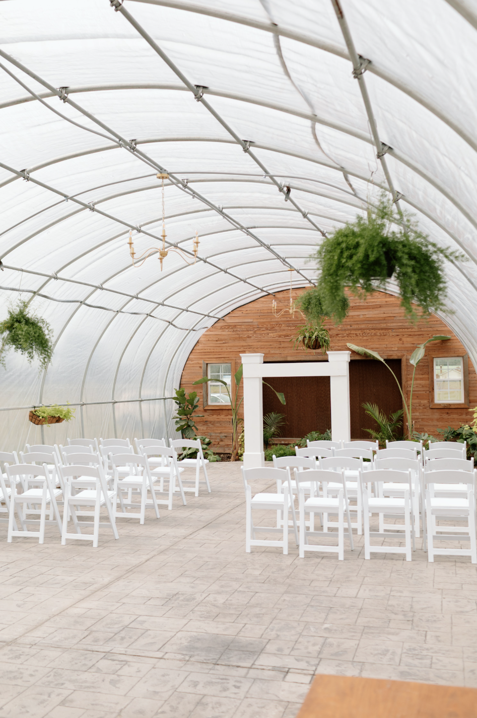Indoor wedding or event space with white chairs arranged in rows, hanging plants, and a wooden wall backdrop with windows and greenery.