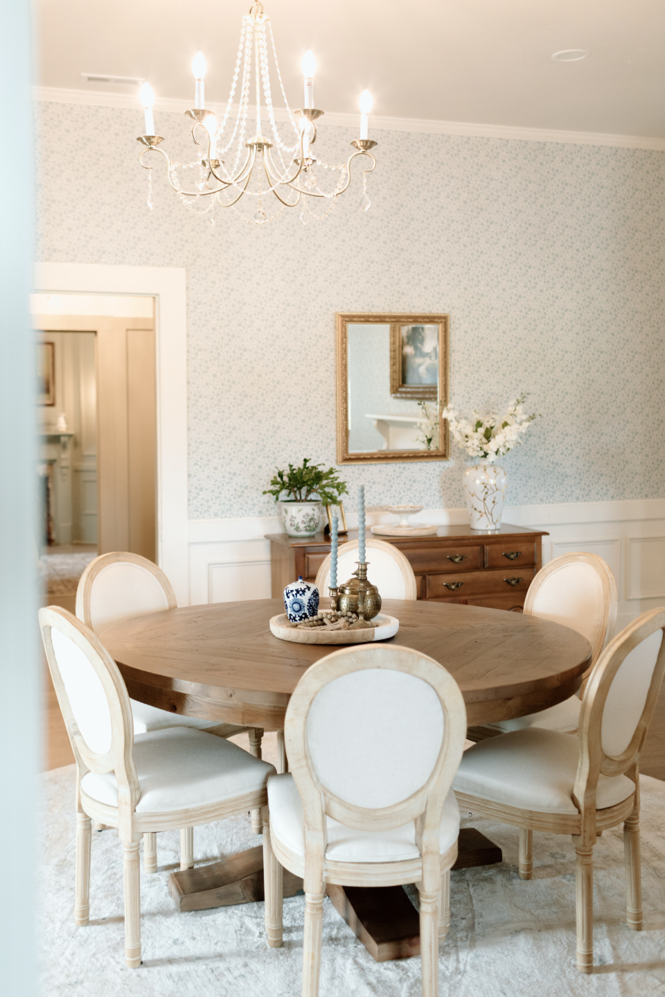 Elegant dining room with a wooden oval table surrounded by six cream-colored upholstered chairs in front of a light blue wall with small floral wallpaper. A chandelier hangs from the ceiling, and a wooden sideboard with a potted plant, a large vase w