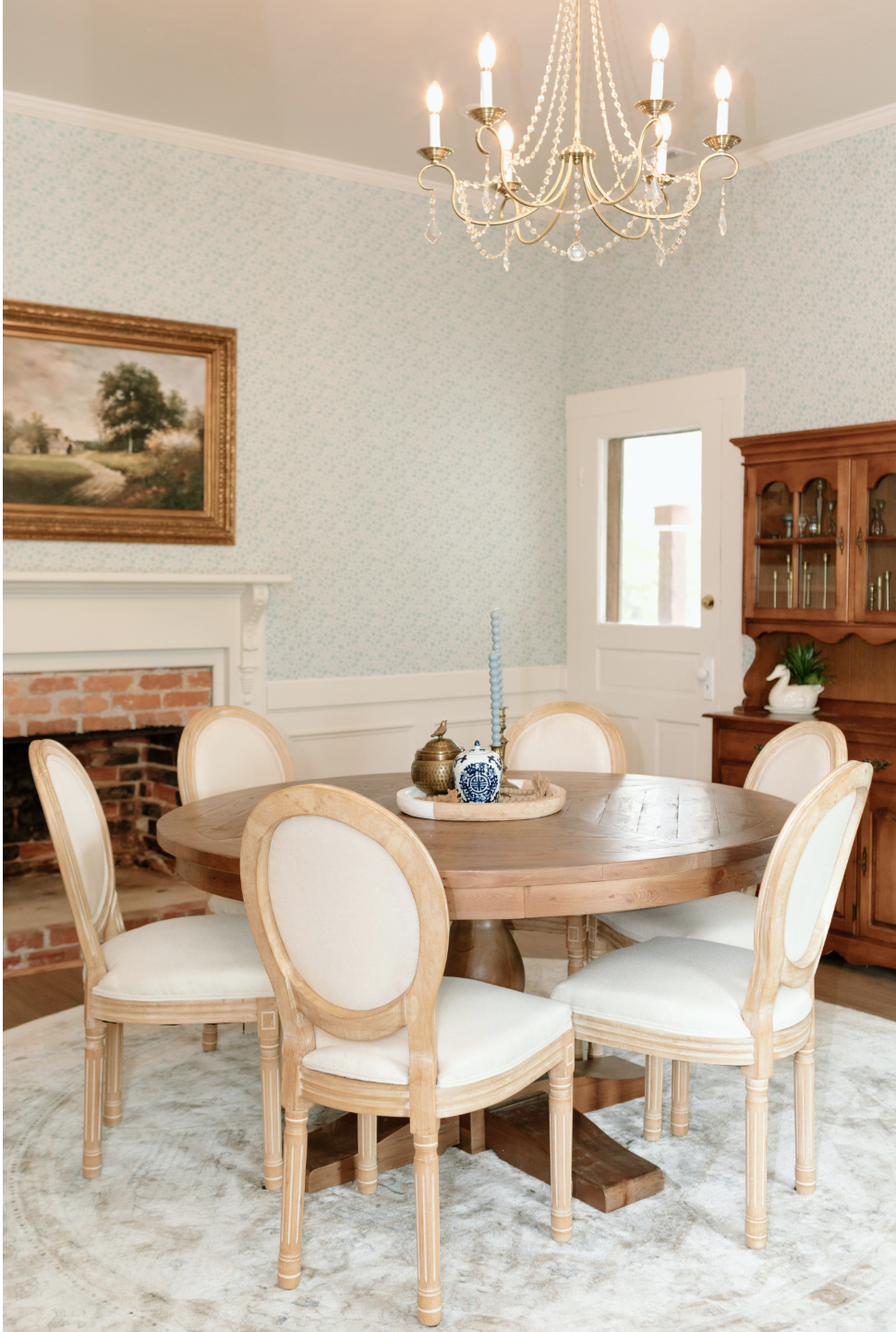 A dining room with a round wooden table, six cream-colored upholstered chairs, a vintage chandelier, a fireplace with a brick interior, a landscape painting on the wall, a wooden hutch with glass doors, and a door leading outside. The table has decor