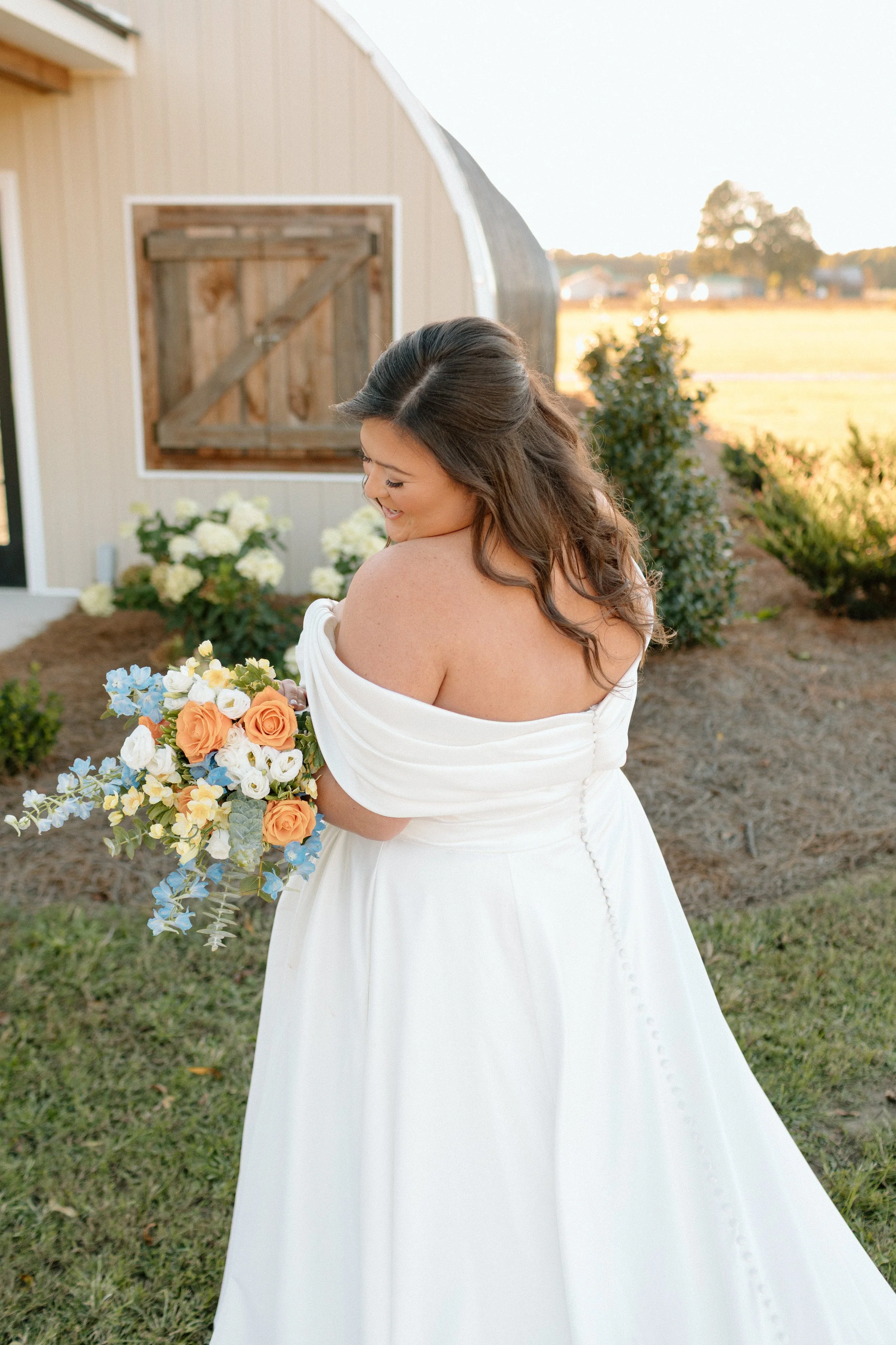 A bride in a white off-the-shoulder wedding dress holding a bouquet of flowers, standing outdoors near a beige barn with a wooden door, during golden hour with trees and a field in the background.
