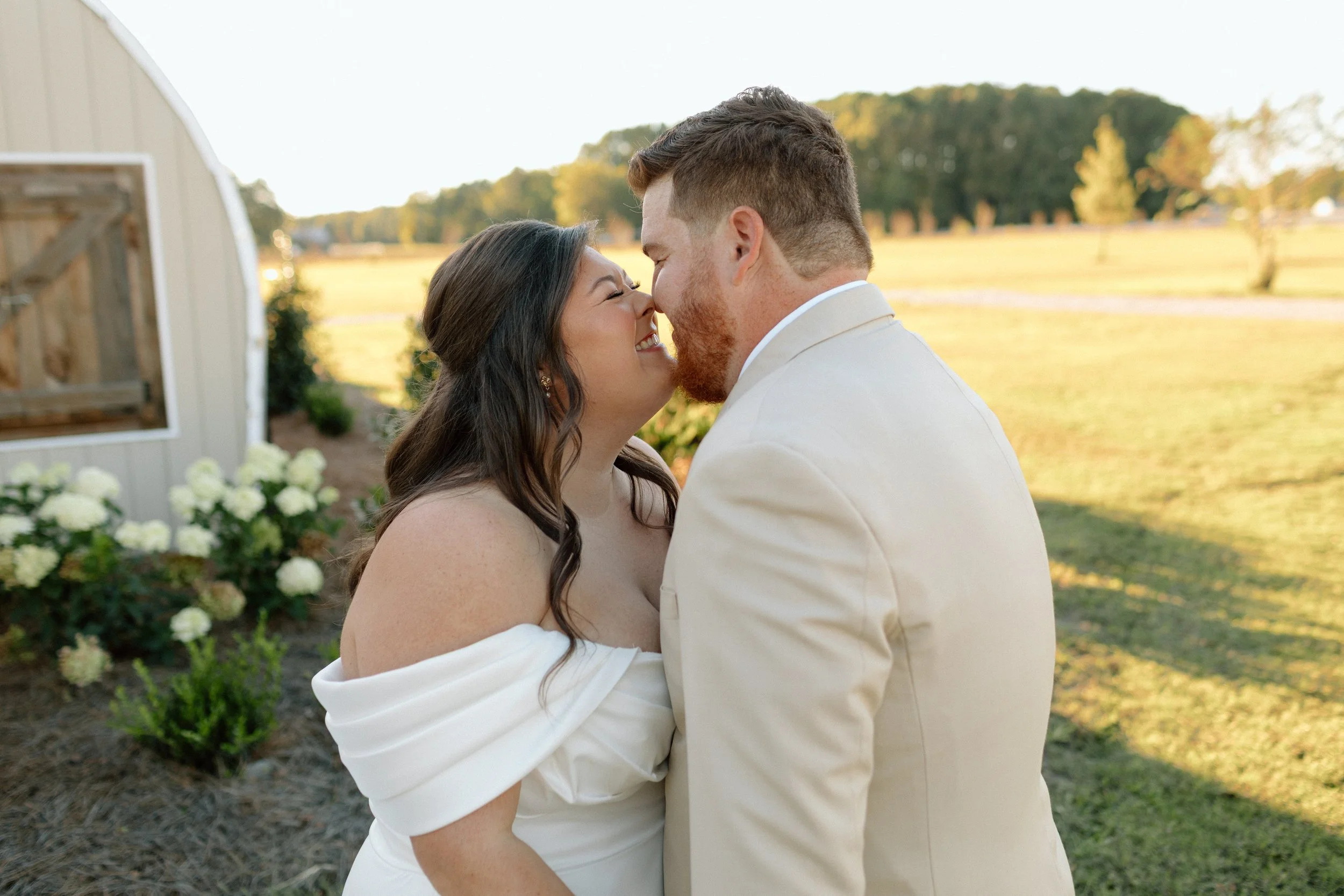 A newlywed couple shares a kiss outdoors in a sunny, rural setting with trees and a field in the background. The woman is wearing a white wedding dress and the man is in a light-colored suit.